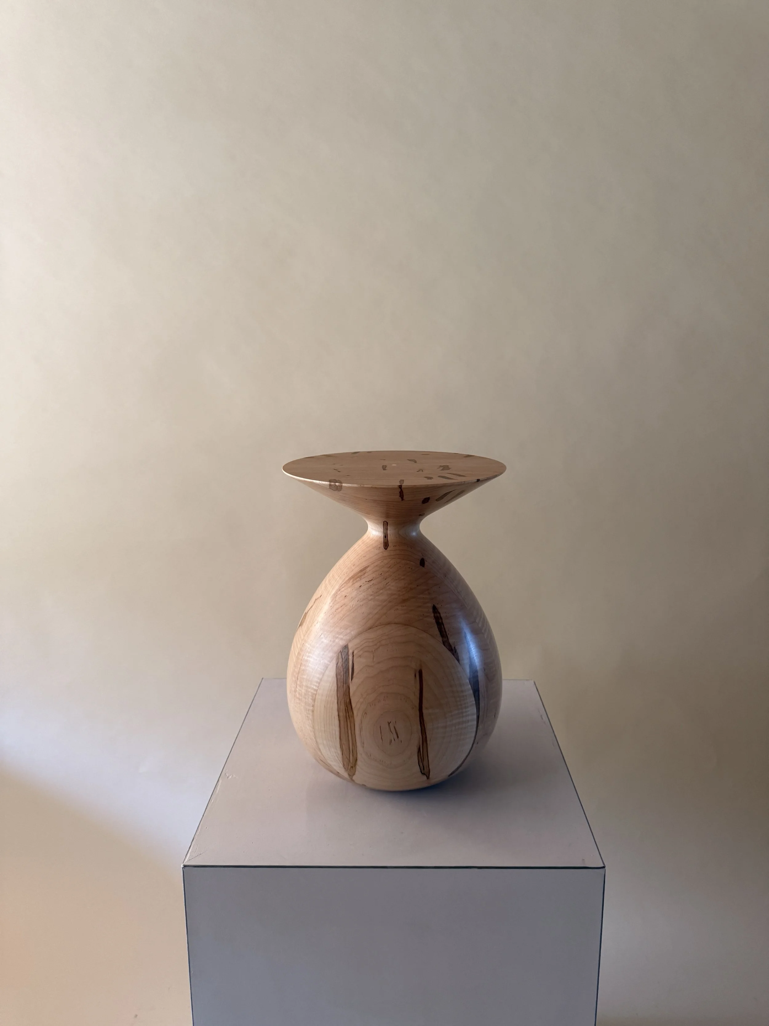 A wooden sculptural art piece with a rounded base and a flat, circular top, displayed on a white pedestal against a neutral wall.