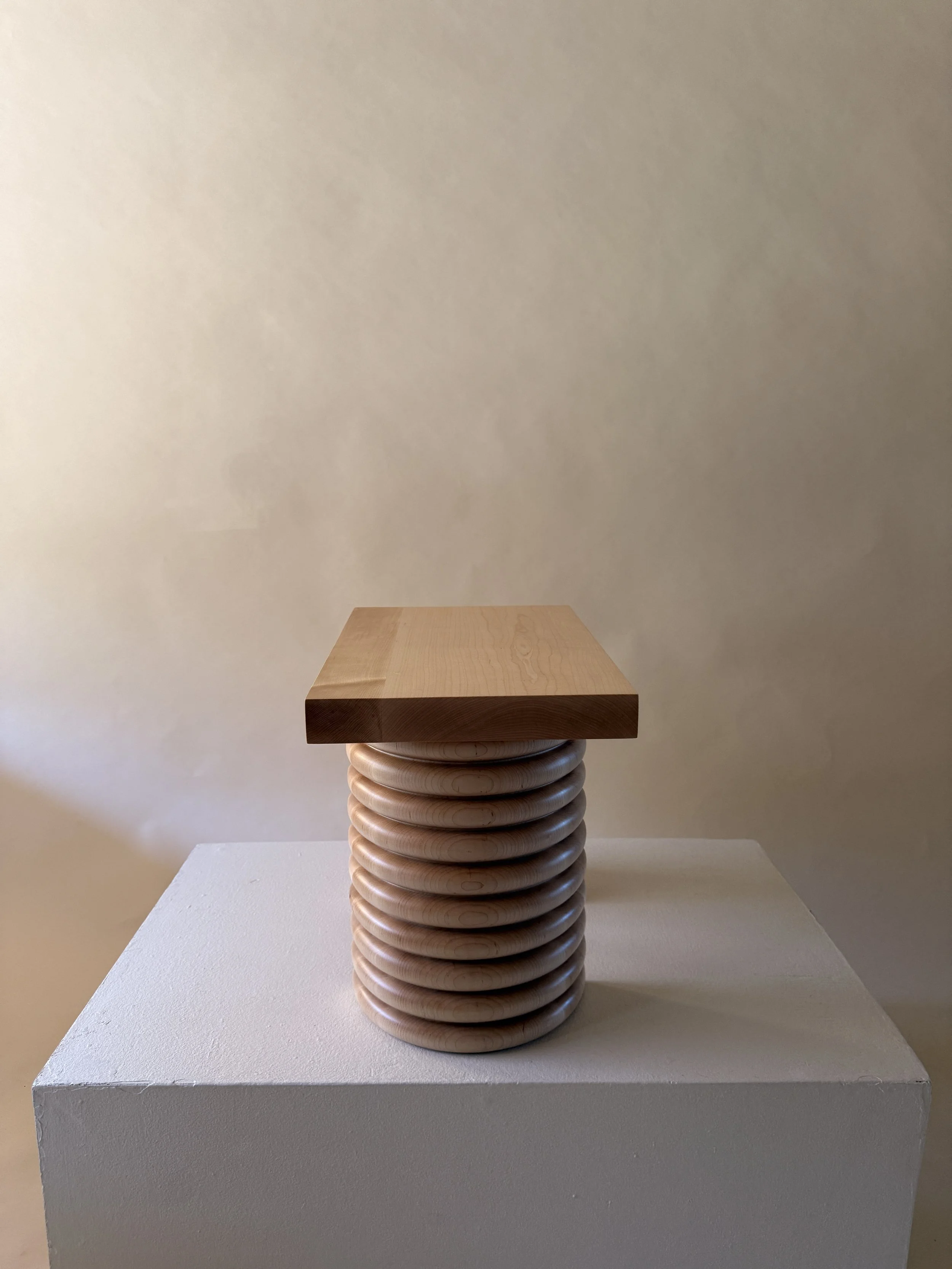 A wooden sculpture featuring a series of concentric rings stacked to form a column, with a flat wooden square on top, set on a white pedestal against a plain, light-colored wall.