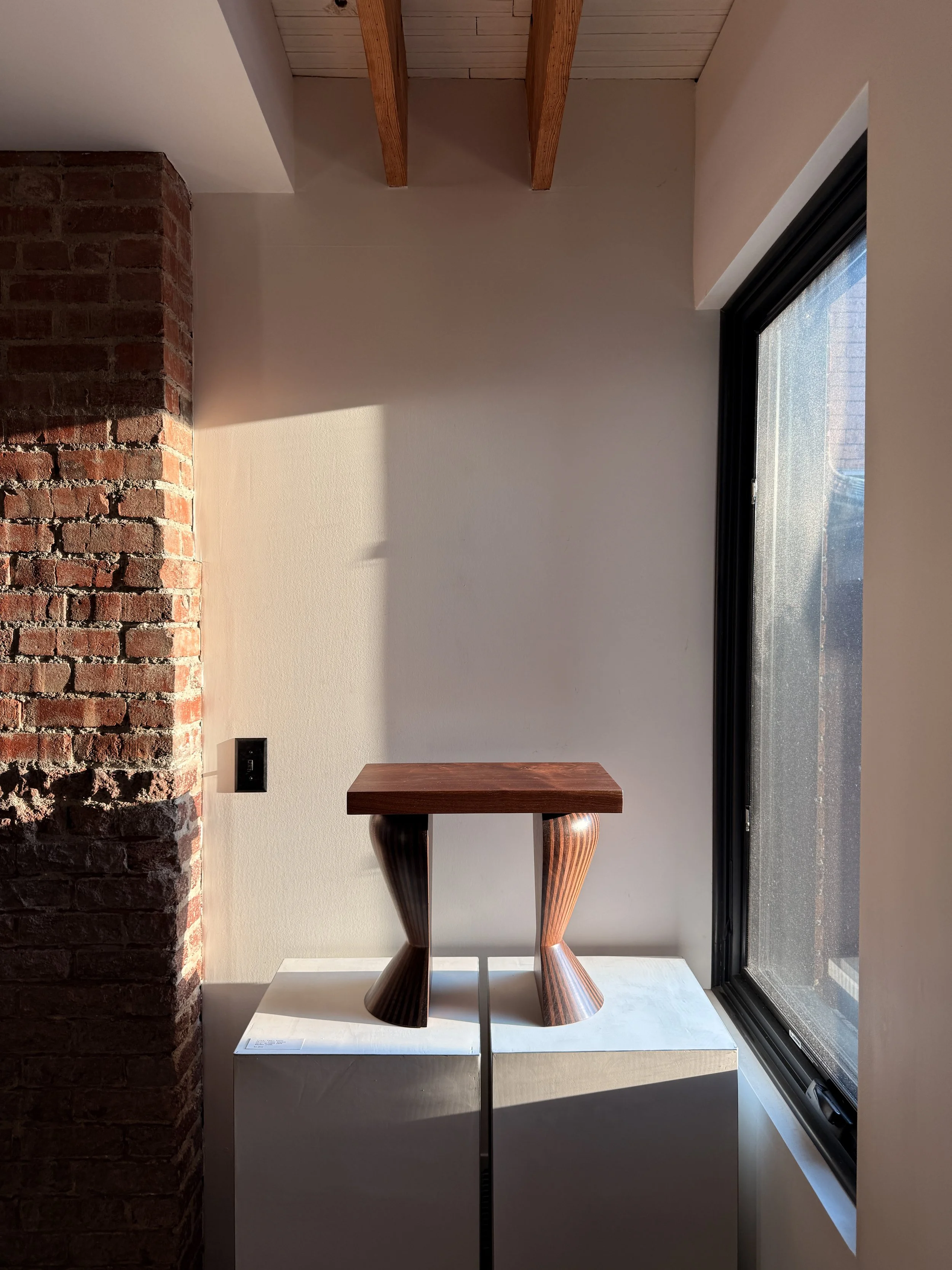 A modern interior space with a brick wall on the left, a beige wall in the center, and a large window on the right, featuring a wooden table with unique curved legs placed on a white base.