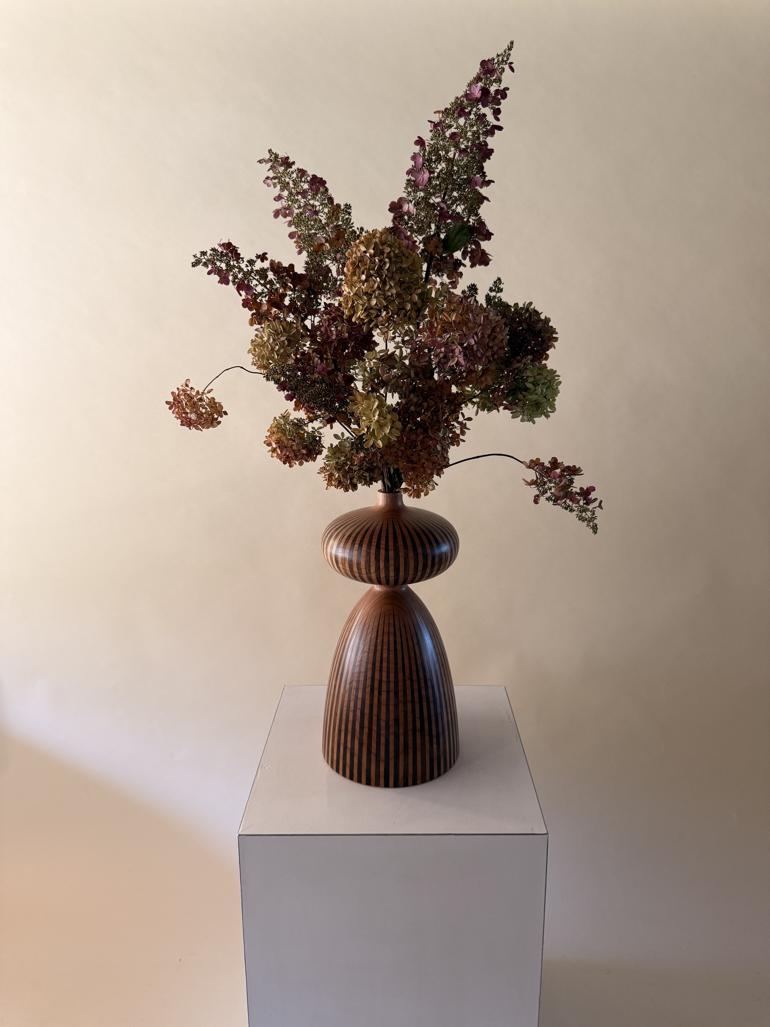 A tall, wooden vase with a striped pattern containing dried pink and beige hydrangea flowers placed on a white pedestal against a plain beige wall.