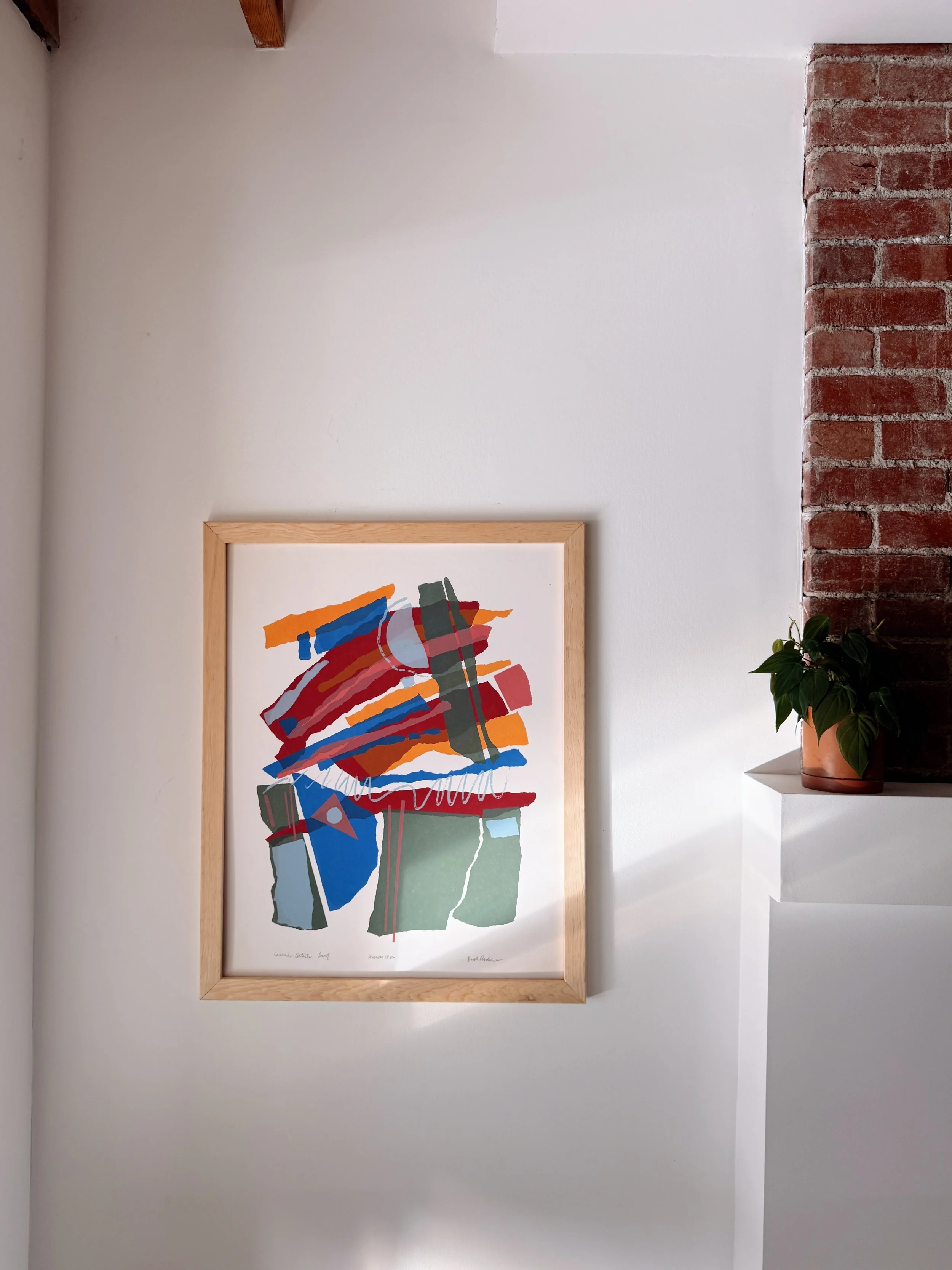 A framed abstract artwork with multicolored brushstrokes in an art gallery, next to a brick wall and a potted plant on a white shelf.