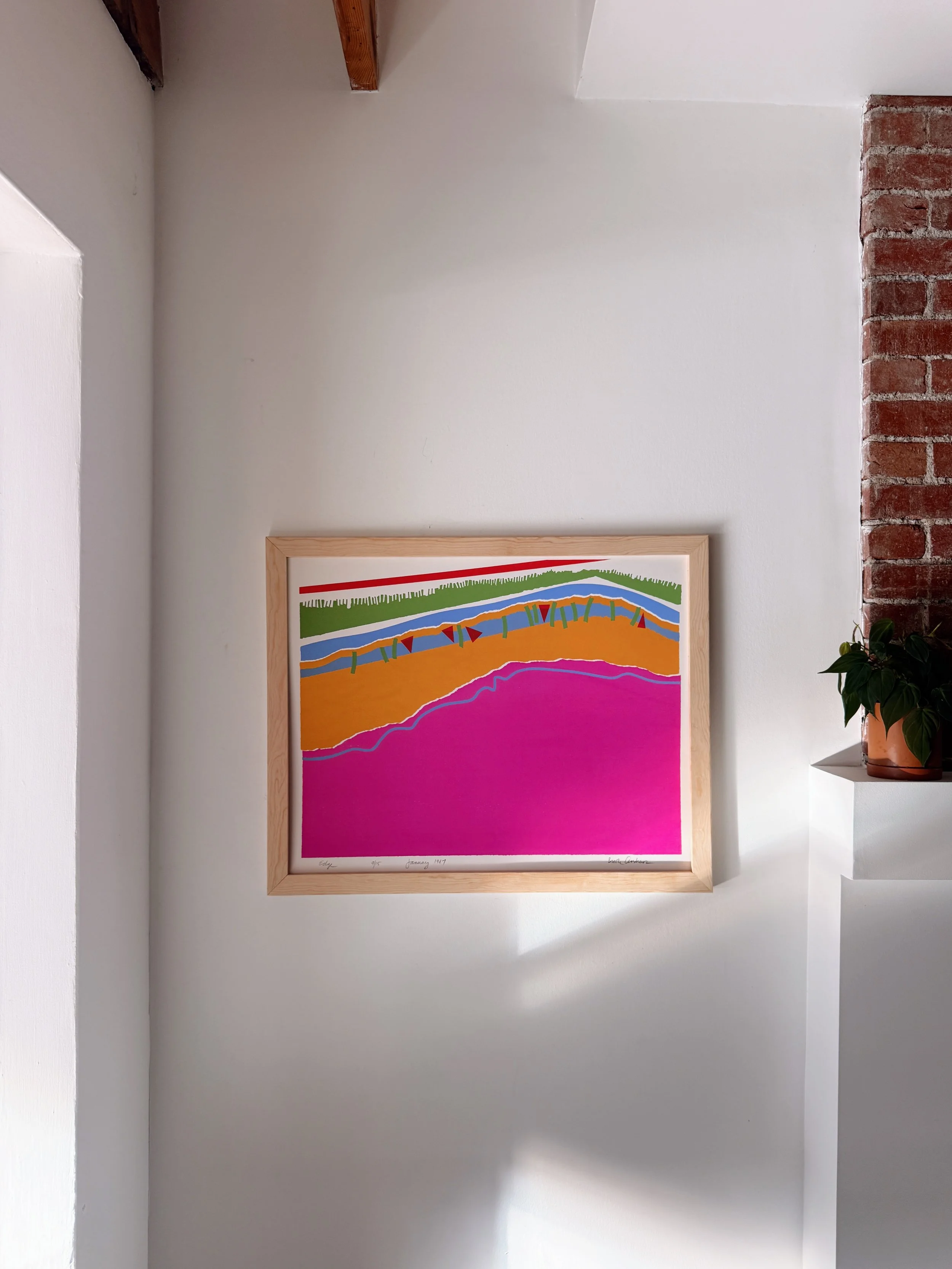 A colorful abstract art piece with horizontal stripes in green, orange, blue, and pink, framed in light wood and displayed on a white wall next to a brick column and a potted plant.