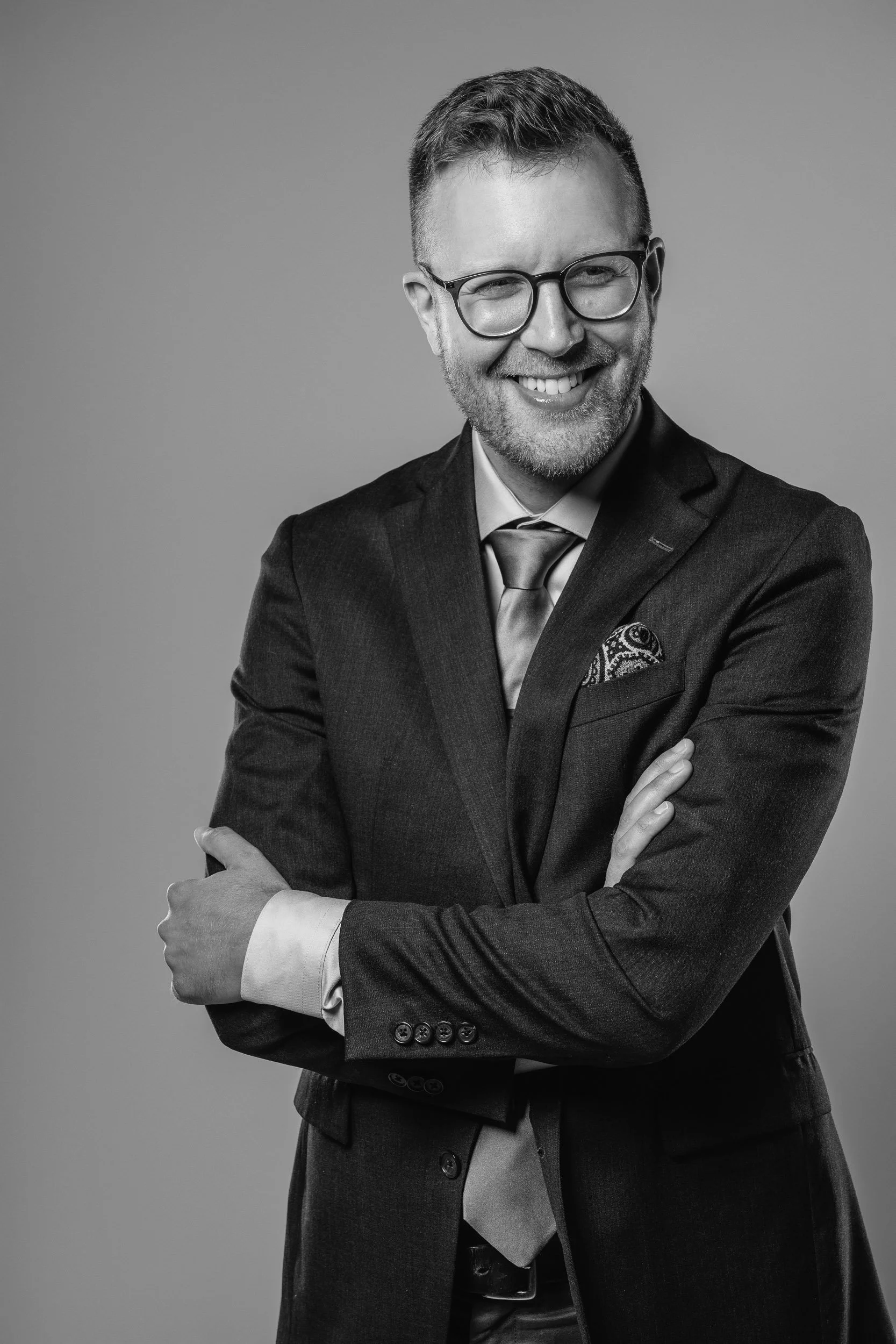 Black and white portrait of a man in a suit smiling, wearing glasses, with arms crossed, against a plain background.