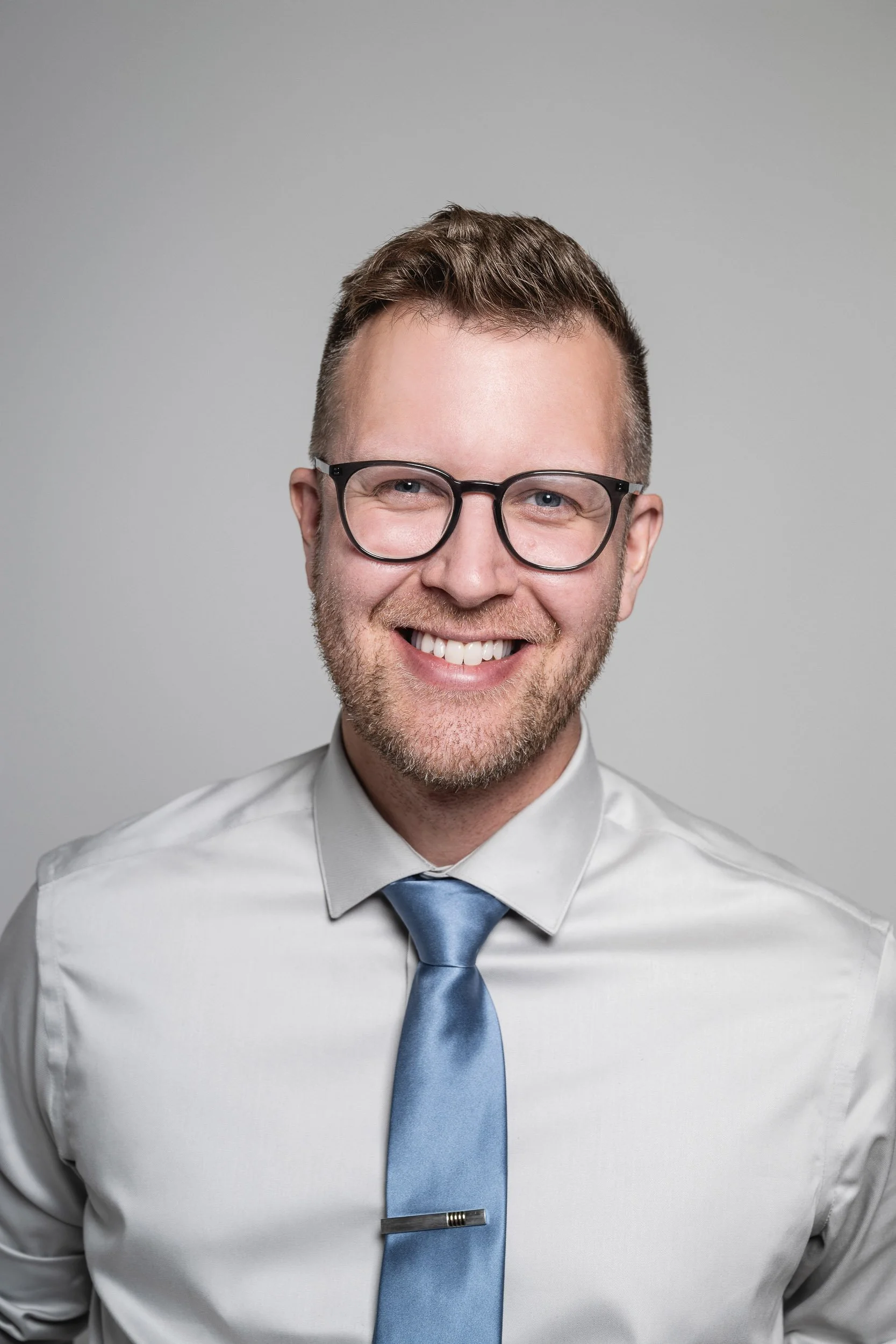 Close-up of a smiling man with glasses, wearing a white shirt and a blue tie against a gray background.