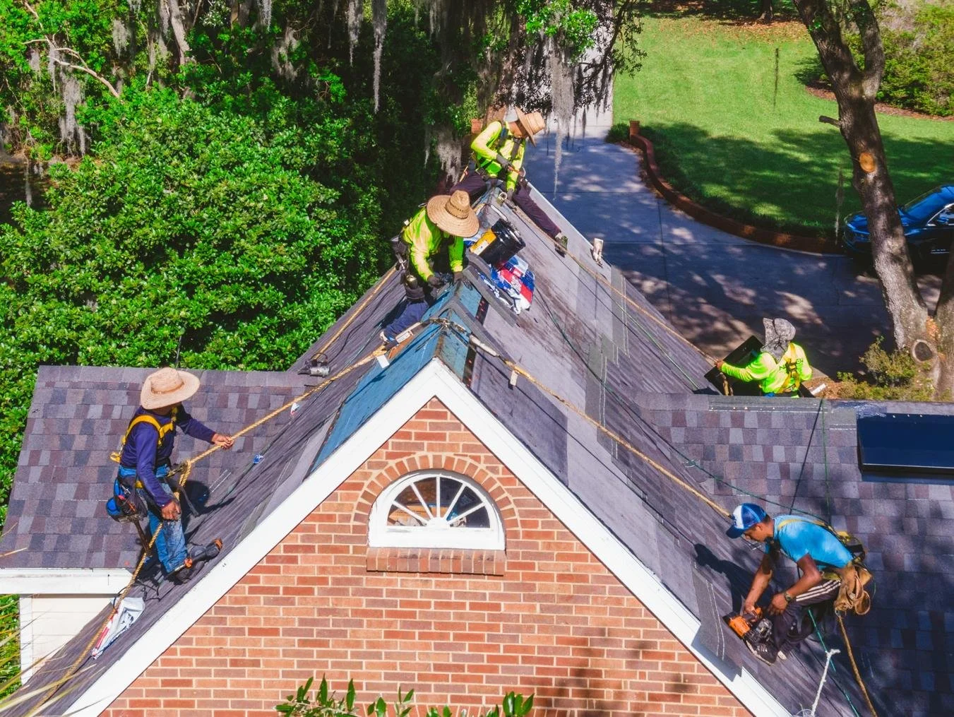 Roofing workers in high-visibility clothing and wide-brimmed hats repairing a house roof. They are using safety harnesses and tools, working from the peak of a gabled brick house with trees and a driveway visible in the background.