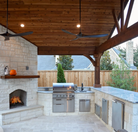 Outdoor kitchen with stone fireplace, stainless steel grill, countertop, and wooden ceiling with ceiling fans.