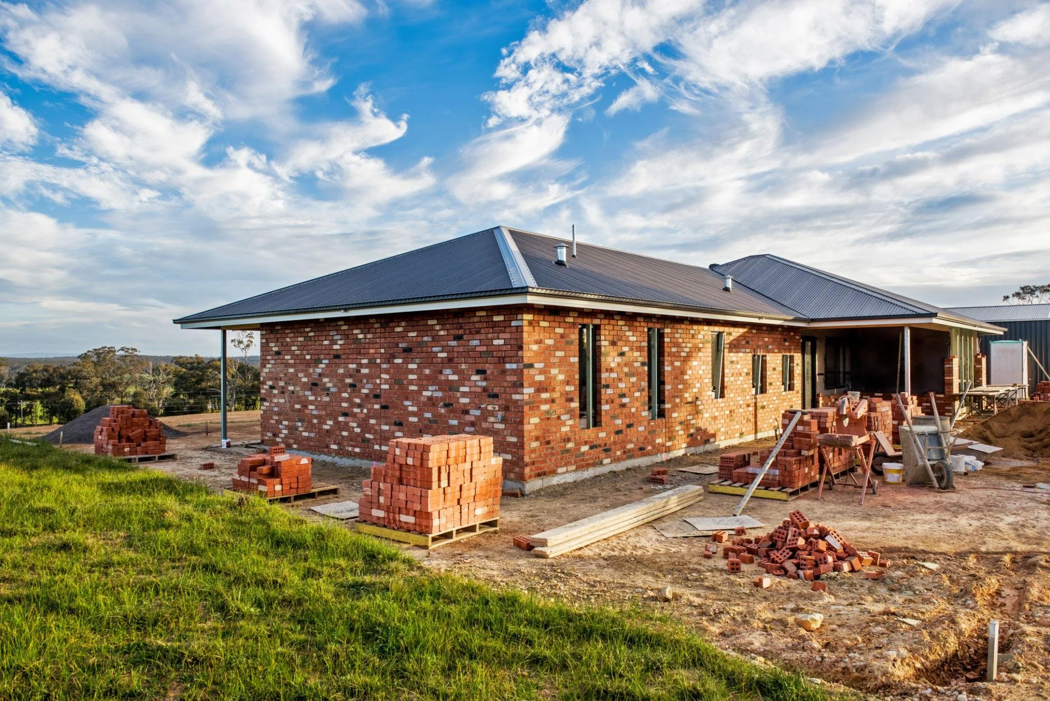 A house under construction with a brick exterior, metal roof, and construction materials and bricks around the site, set against a landscape with trees and a cloudy sky.