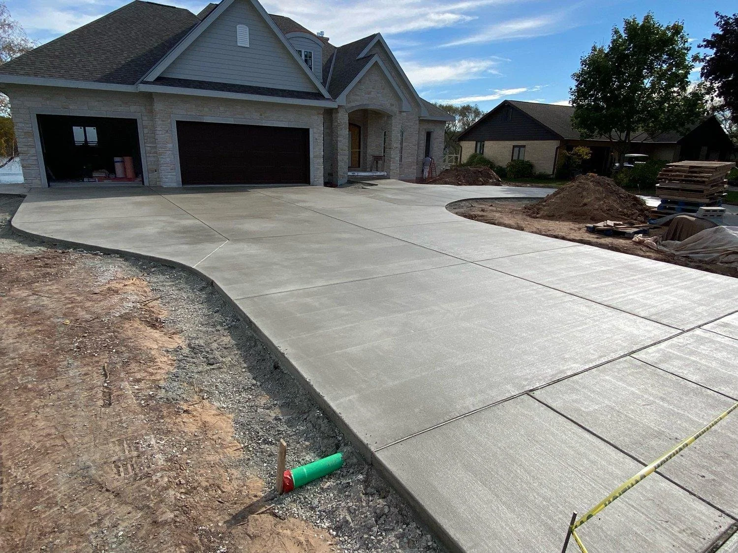 Newly poured concrete driveway in front of a house under construction, with construction materials and dirt piles nearby.