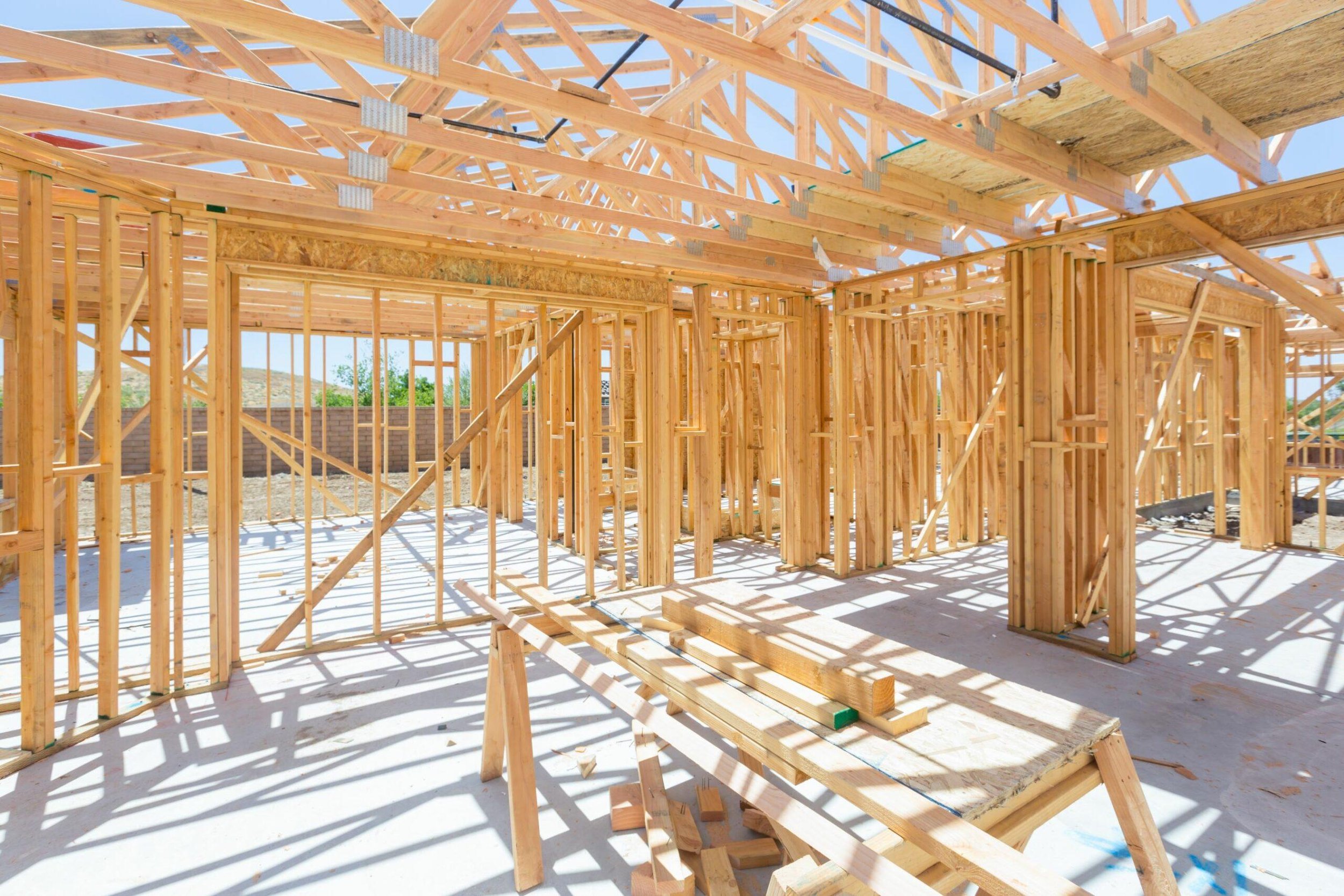 Wooden framing structure of a house under construction with partly assembled roof trusses and framing walls, shadows cast on the concrete floor, and a clear blue sky in the background.