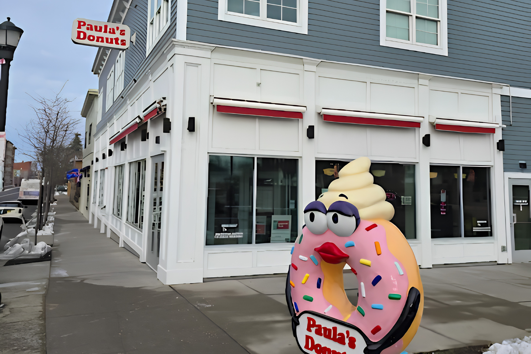 A colorful cupcake-shaped character with eyes, lips, sprinkles on frosting, and a sign reading 'Paula's Donuts' outside a donut shop with blue siding and large windows.