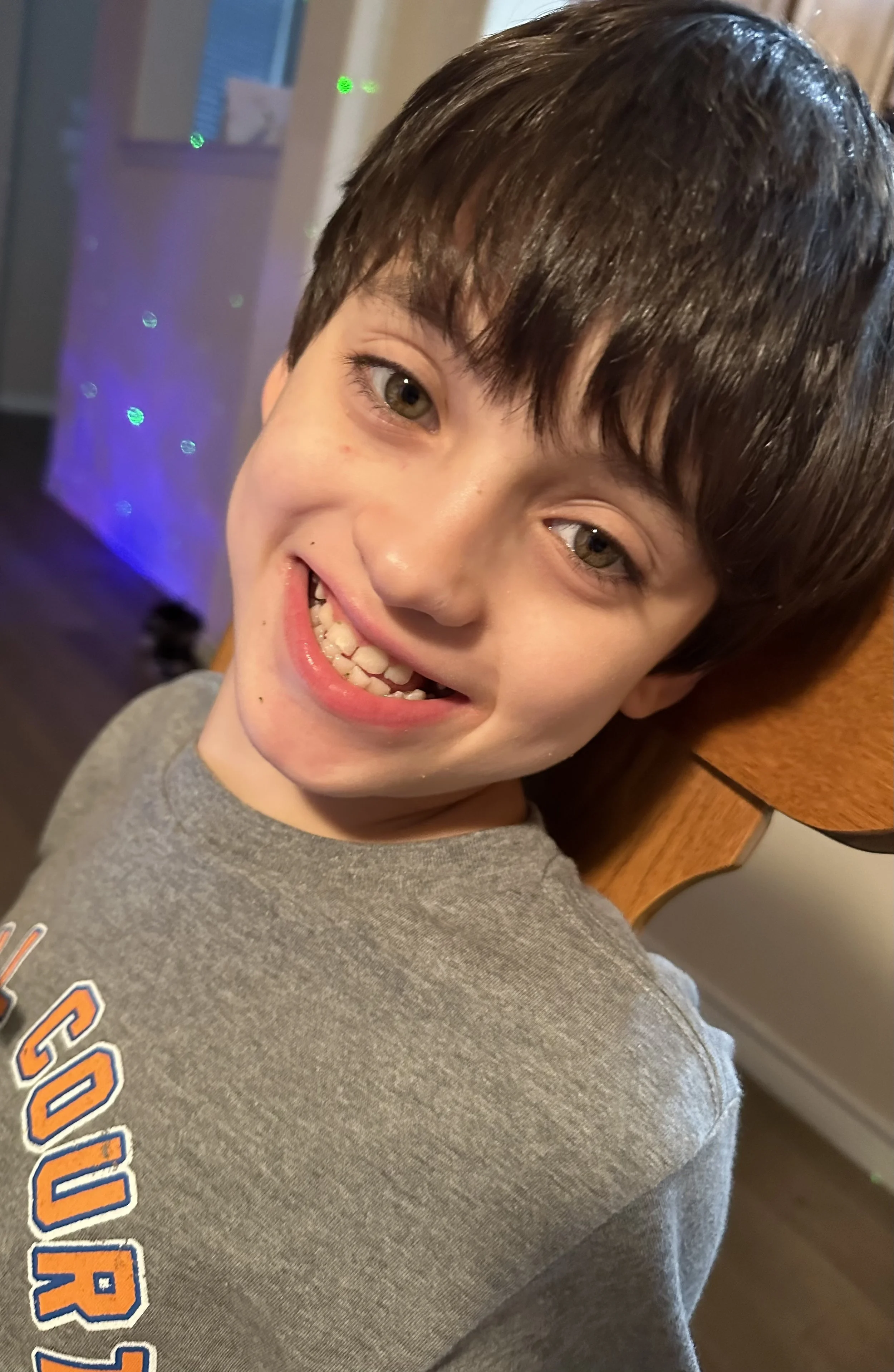 Close-up of a smiling young boy with brown hair and green eyes, wearing a grey shirt with orange and blue lettering, standing indoors near a wooden table.