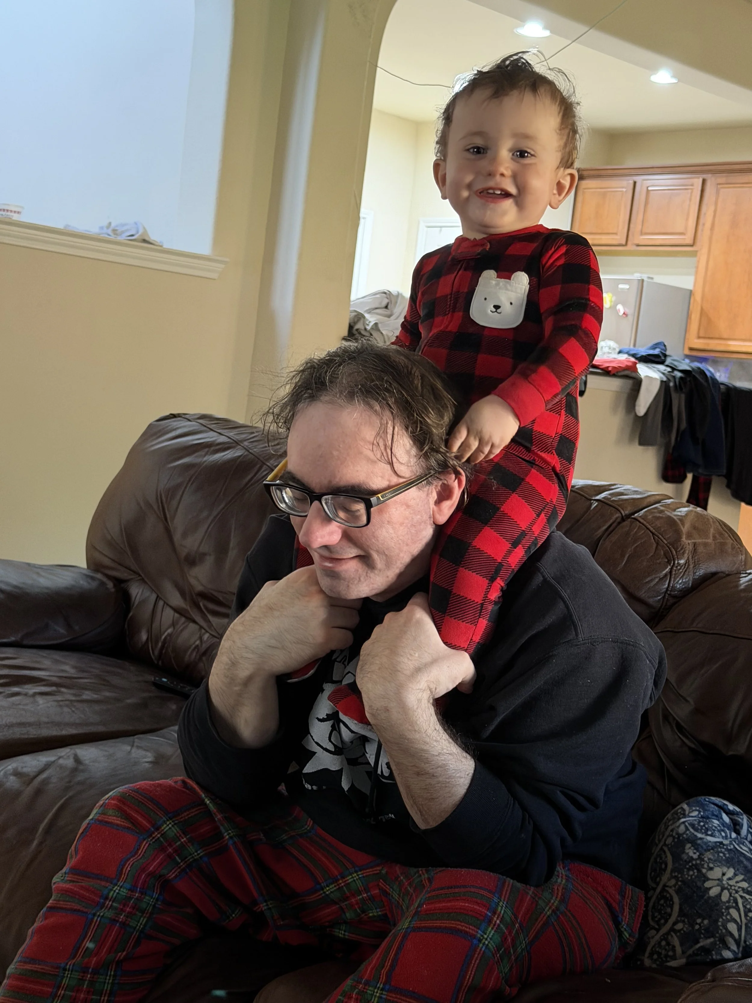 A young child with curly hair, wearing red and black plaid pajamas with a bear patch, sitting on an adult's shoulders while smiling at the camera. The adult is seated on a brown leather couch, wearing glasses and a dark shirt. The background shows a kitchen with wooden cabinets and laundry on the counter.