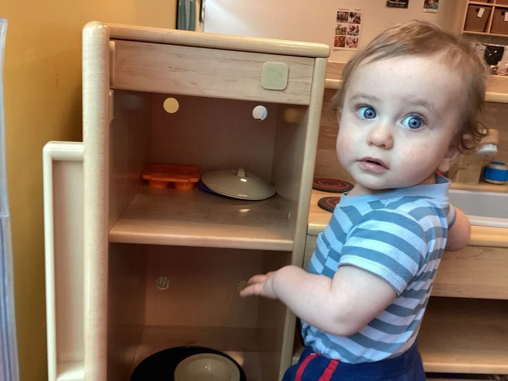A young child with blue eyes and curly hair stands next to a wooden kitchen playset, reaching towards the lower shelf. The child is wearing a blue and gray striped shirt.