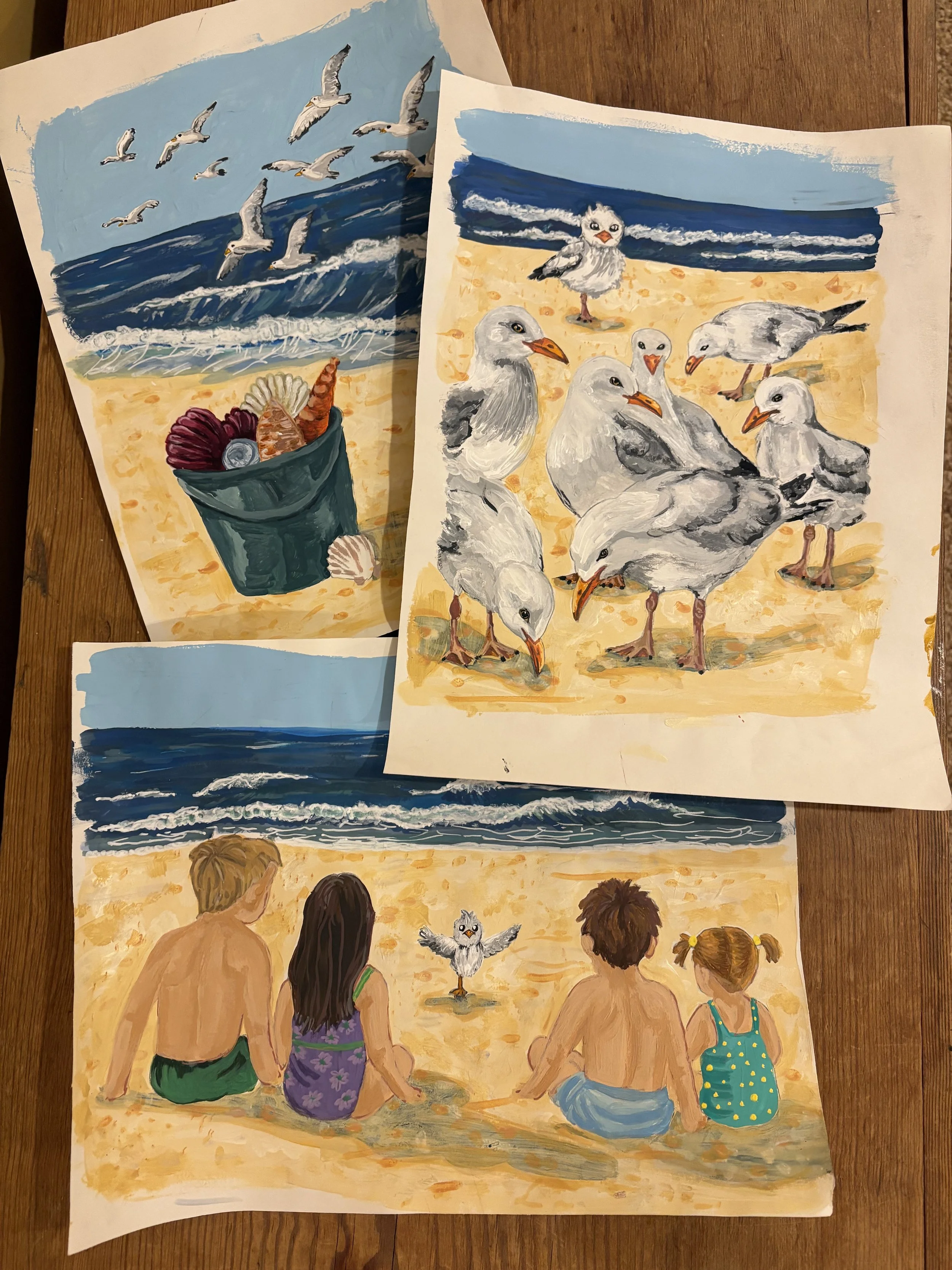 Four children, three girls and a boy, sitting on a sandy beach watching seagulls and other birds. Two of the girls are in a green dress and purple dress with floral patterns, the boy is shirtless in blue shorts, and the girl on the right wears a turquoise swimsuit with yellow dots. In the background, there are three seagulls and other seabirds flying near the ocean waves.