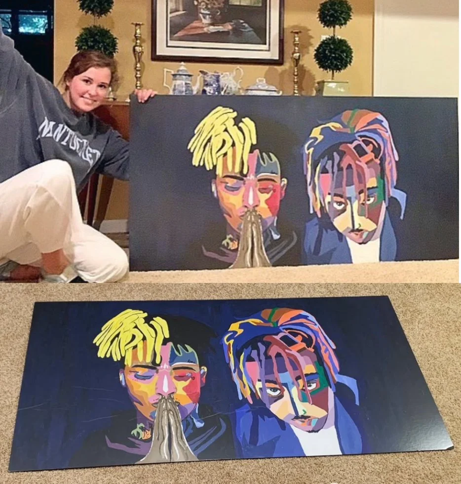 A young woman smiling and kneeling next to two colorful portrait paintings of two men, with the paintings on the carpet and one on an easel or surface behind her. The paintings depict two men with unique, colorful hairstyles and expressions, in a stylized, abstract art style.