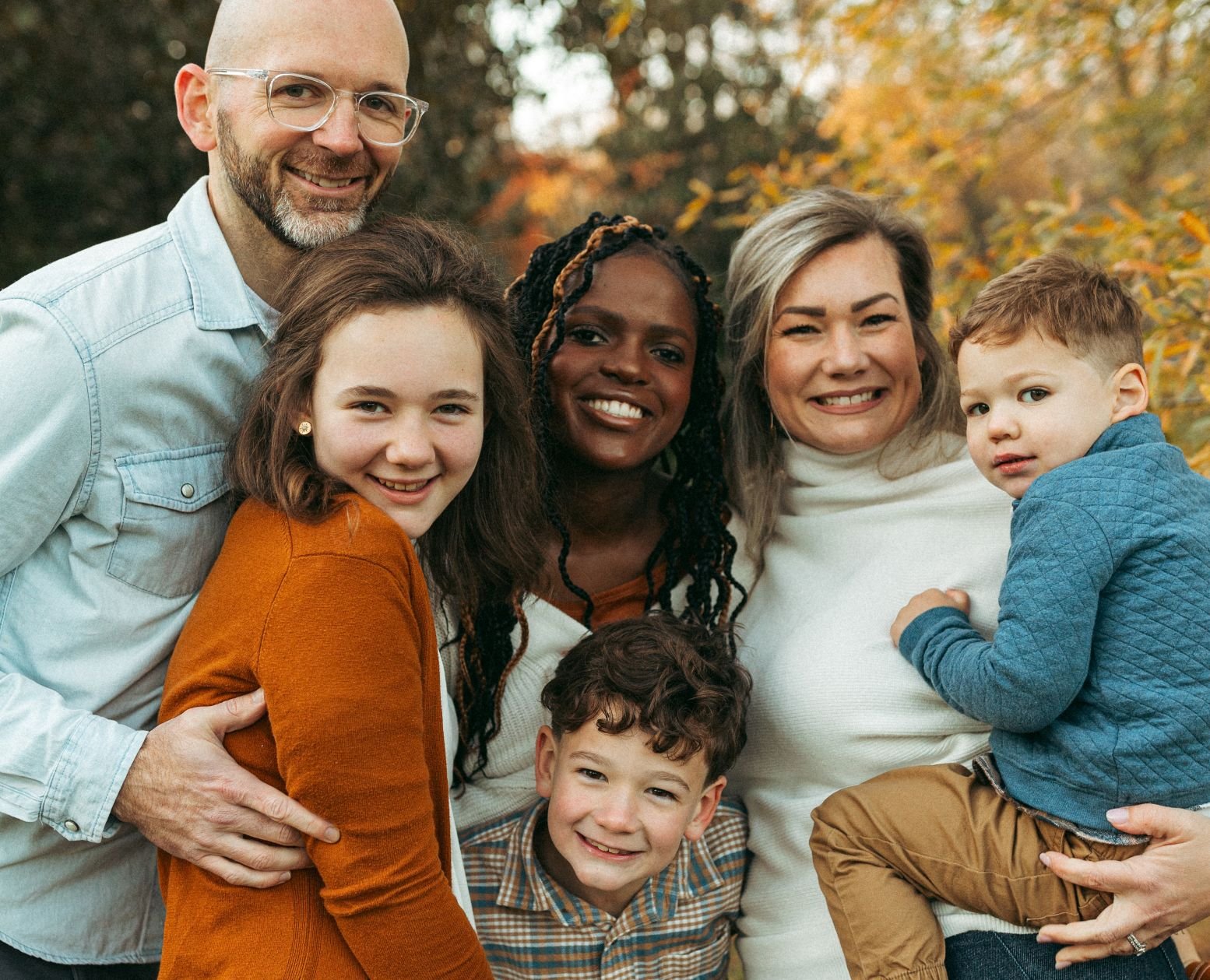 A group of six people smiling outdoors in autumn, including two men, two women, and two children, one girl and one boy, surrounded by fall foliage.