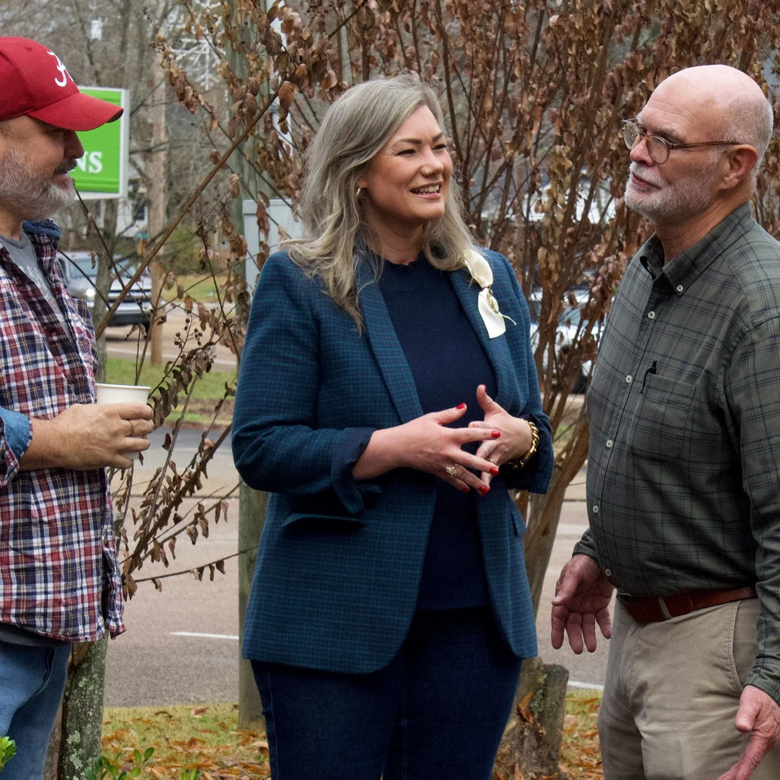 Three people are engaged in conversation outdoors near a tree with brown leaves. One woman and two men are visible. The woman is smiling, wearing a blue checkered blazer, black shirt, and jewelry, and has blonde hair. The man on her right is wearing glasses, a plaid shirt, and khaki pants. The man on her left is wearing a red cap, plaid shirt, and holding a cup.