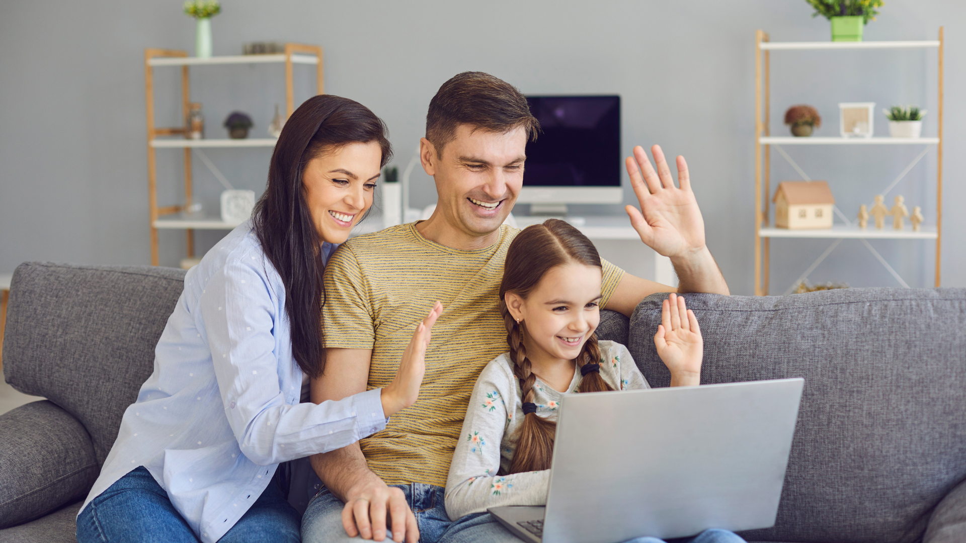 Family of three using a laptop on a couch, smiling and waving at the screen in a modern, cozy living room.