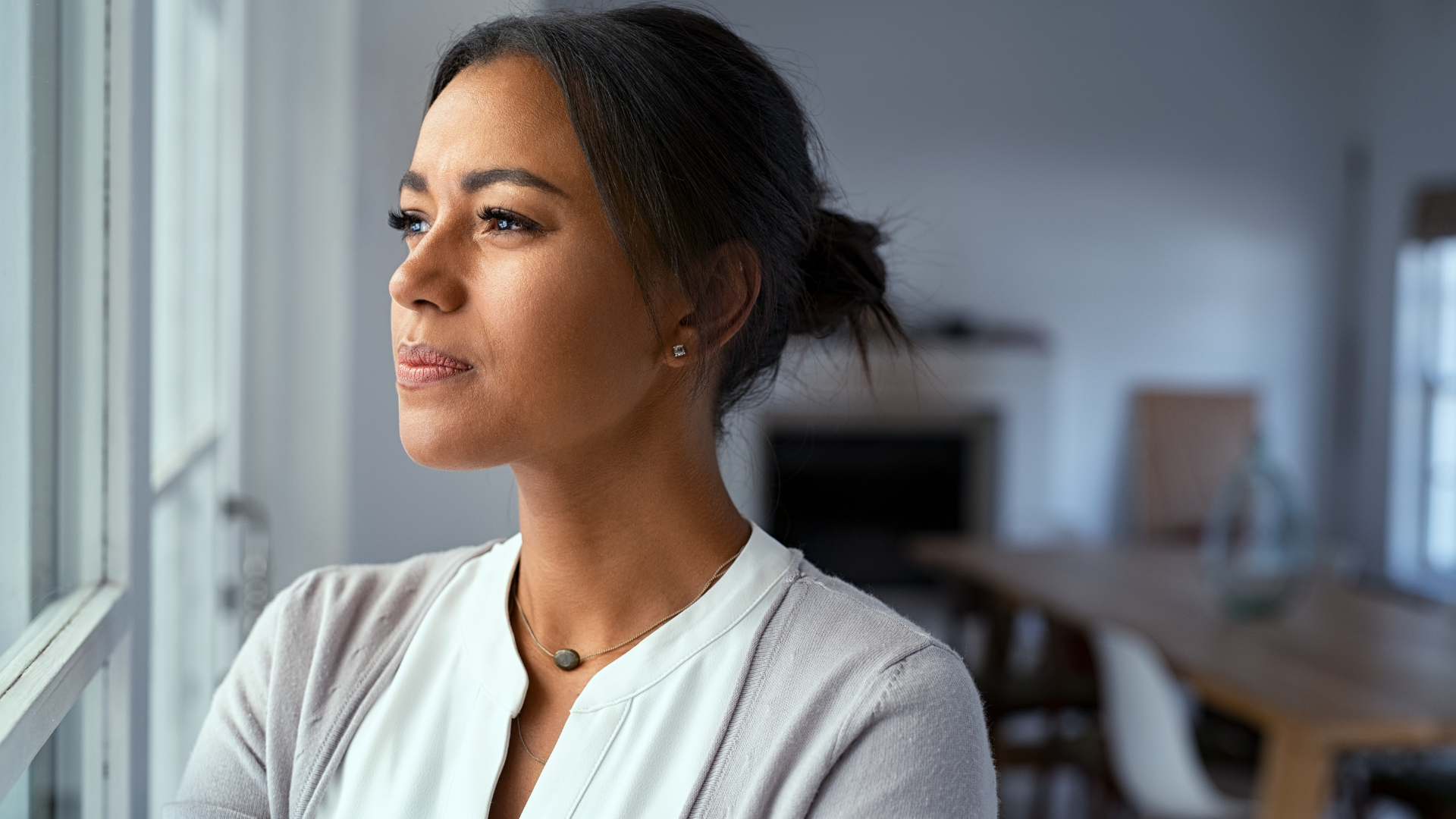 Thoughtful woman looking out window in home setting, wearing a light-colored blouse and cardigan, with hair in a bun.