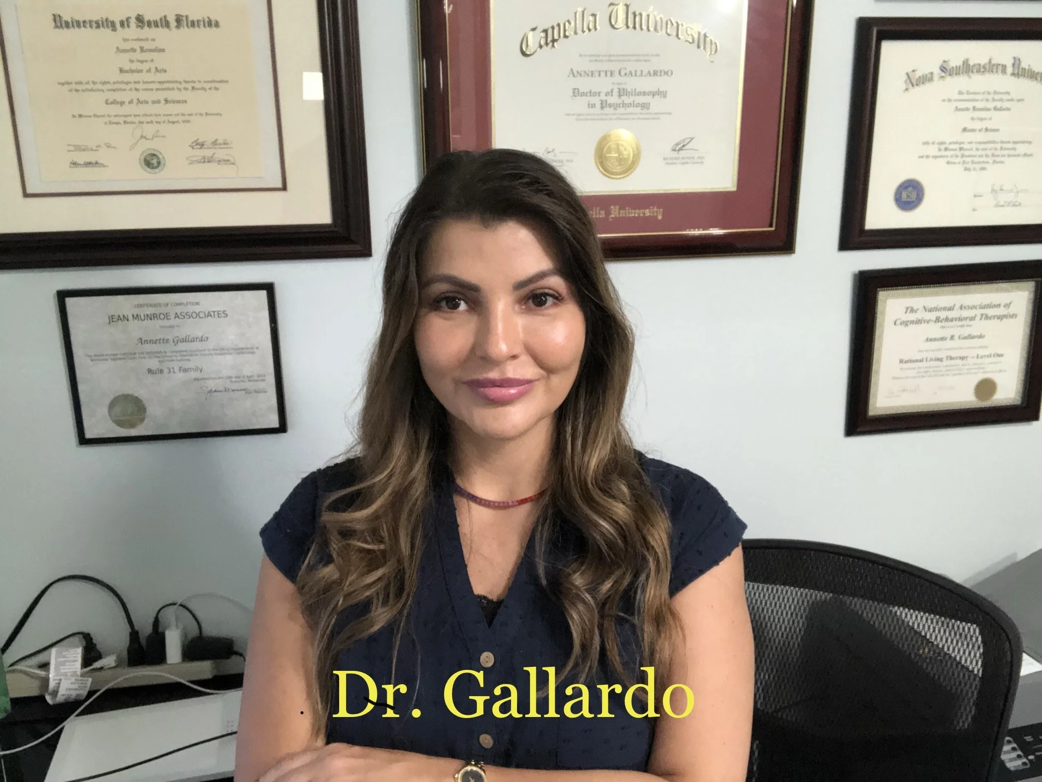 A woman standing in front of a wall displaying multiple educational certificates and diplomas. She has long hair and is wearing a dark blouse. The certificates are framed and arranged neatly on the wall behind her. One digital display shows the text "Dr. Gallardo."