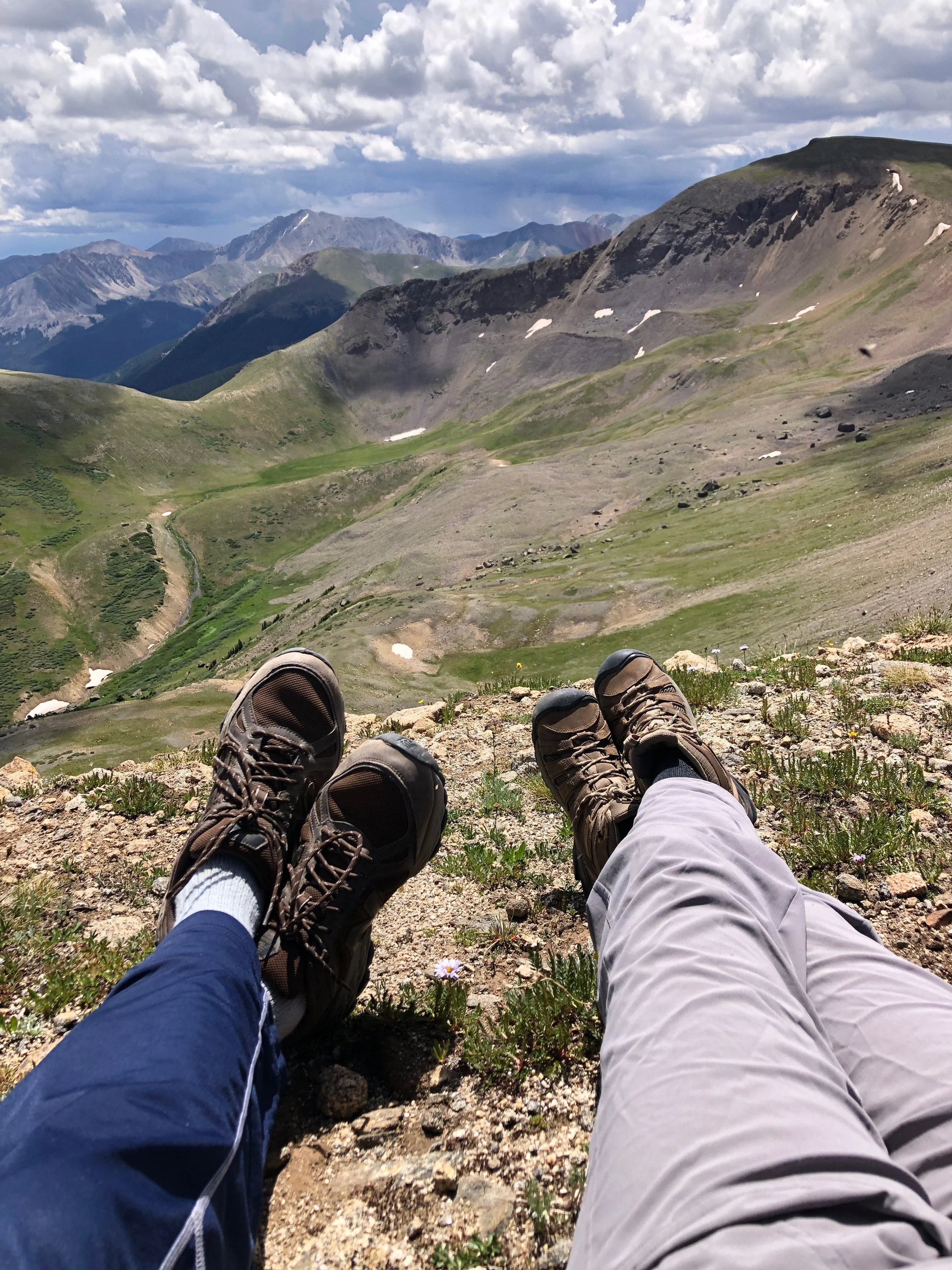 Two hikers' legs and feet wearing hiking boots and pants, resting on a grassy mountain slope with a vast mountain landscape and cloudy sky in the background.
