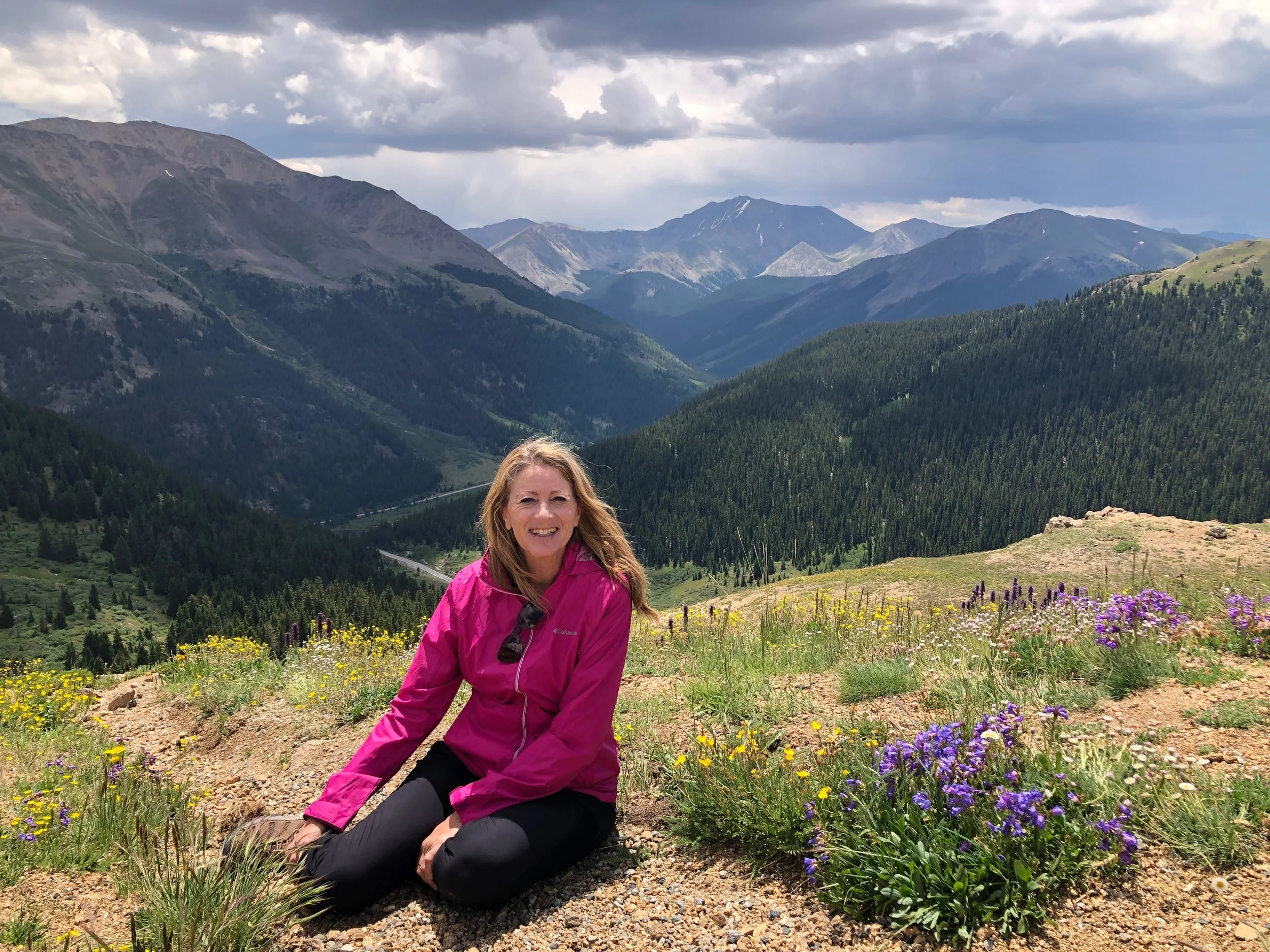 A woman wearing a pink jacket and black pants sitting on rocky ground surrounded by wildflowers with mountain ranges and a cloudy sky in the background.