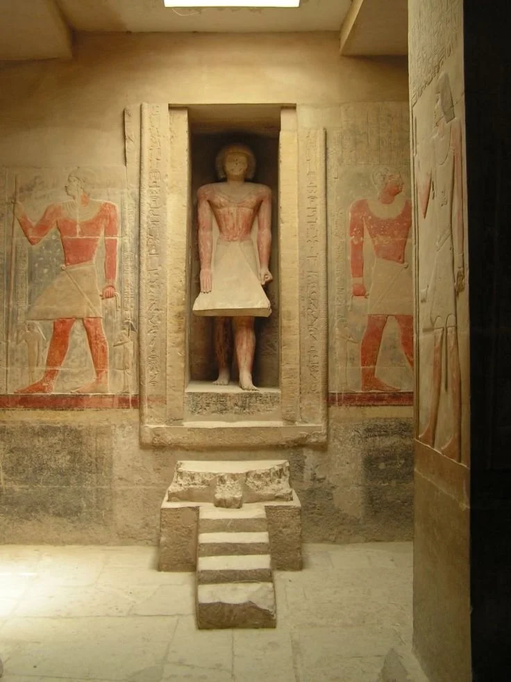 Ancient Egyptian sculpture of a standing figure, flanked by wall paintings of two other figures with hieroglyphics, in a museum display.