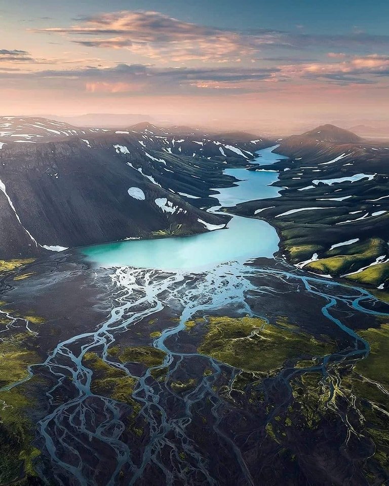 Aerial view of a volcanic landscape with a large turquoise crater lake, dark mountains with patches of snow, and a network of glacial streams flowing through green mossy terrain, under a pastel-colored sky at sunset.