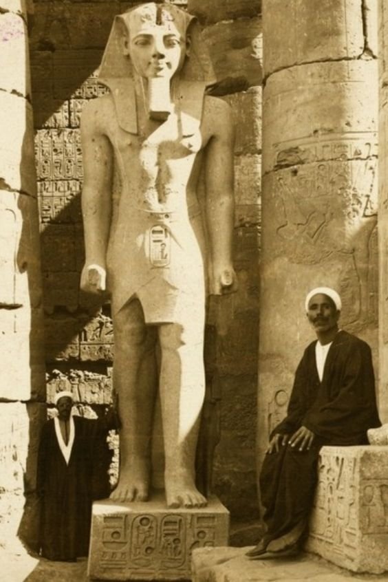 A sepia-toned photograph of two men dressed in traditional Middle Eastern attire, standing near a large ancient Egyptian statue of a pharaoh.