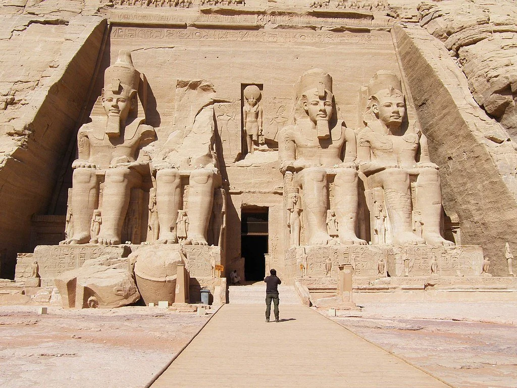Large ancient Egyptian statues of pharaohs at the entrance of a temple, carved from stone with hieroglyphics, located in a desert area.