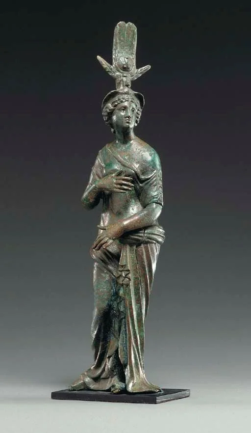 Ancient bronze sculpture of a woman with a headdress, dressed in traditional clothing, standing with one hand on her chest and the other resting on her skirt.