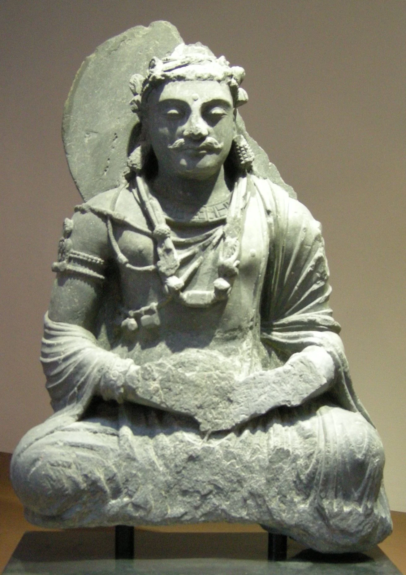 Ancient stone sculpture of a seated figure with traditional attire and jewelry, possibly of historical or cultural significance.
