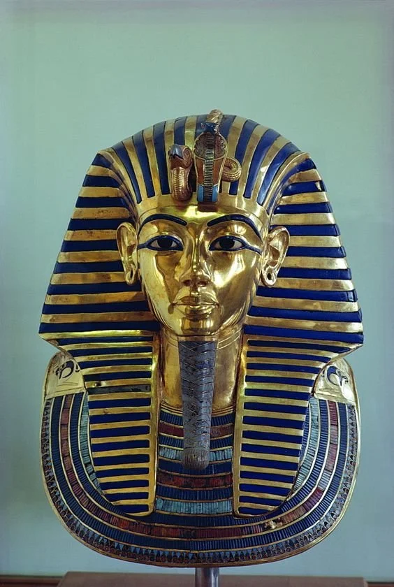A golden death mask of an ancient Egyptian pharaoh with a striped blue and gold nemes headdress and a decorative collar.