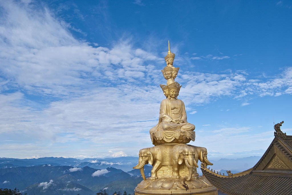 A golden statue of a seated Buddha with multiple faces stacked above, situated outdoors against a blue sky with clouds, mountains, and traditional tiled temple roofs nearby.