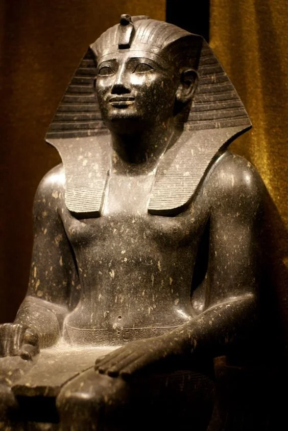 Ancient Egyptian statue of a pharaoh with striped nemes headdress, sitting with hands resting on lap, in dark stone with visible wear.