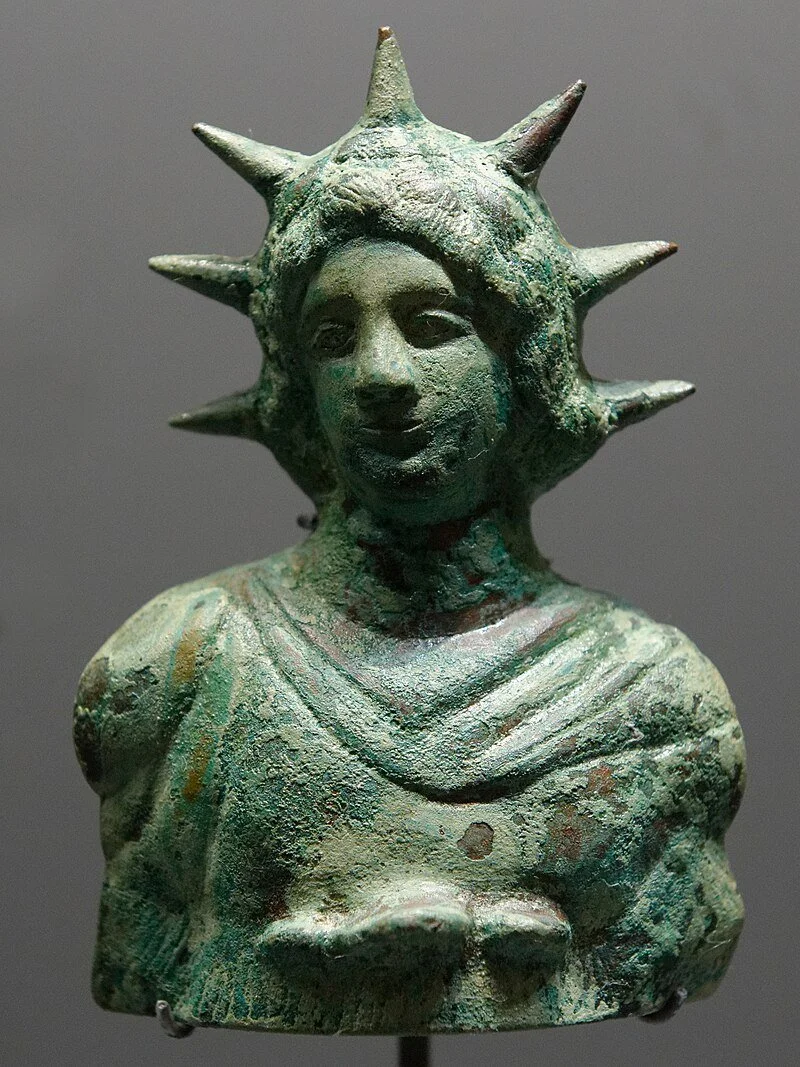 A bronze bust sculpture of the Statue of Liberty's head with signature spiked crown, showing weathered and aged green patina.