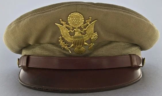 A beige military cap with a gold U.S. emblem featuring an eagle atop a shield, set against a plain background.