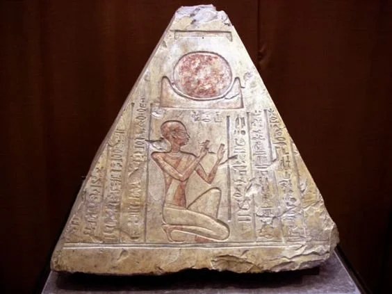 Ancient Egyptian carved stone with hieroglyphs and figure, featuring a seated person with one arm raised and a sun disk above.