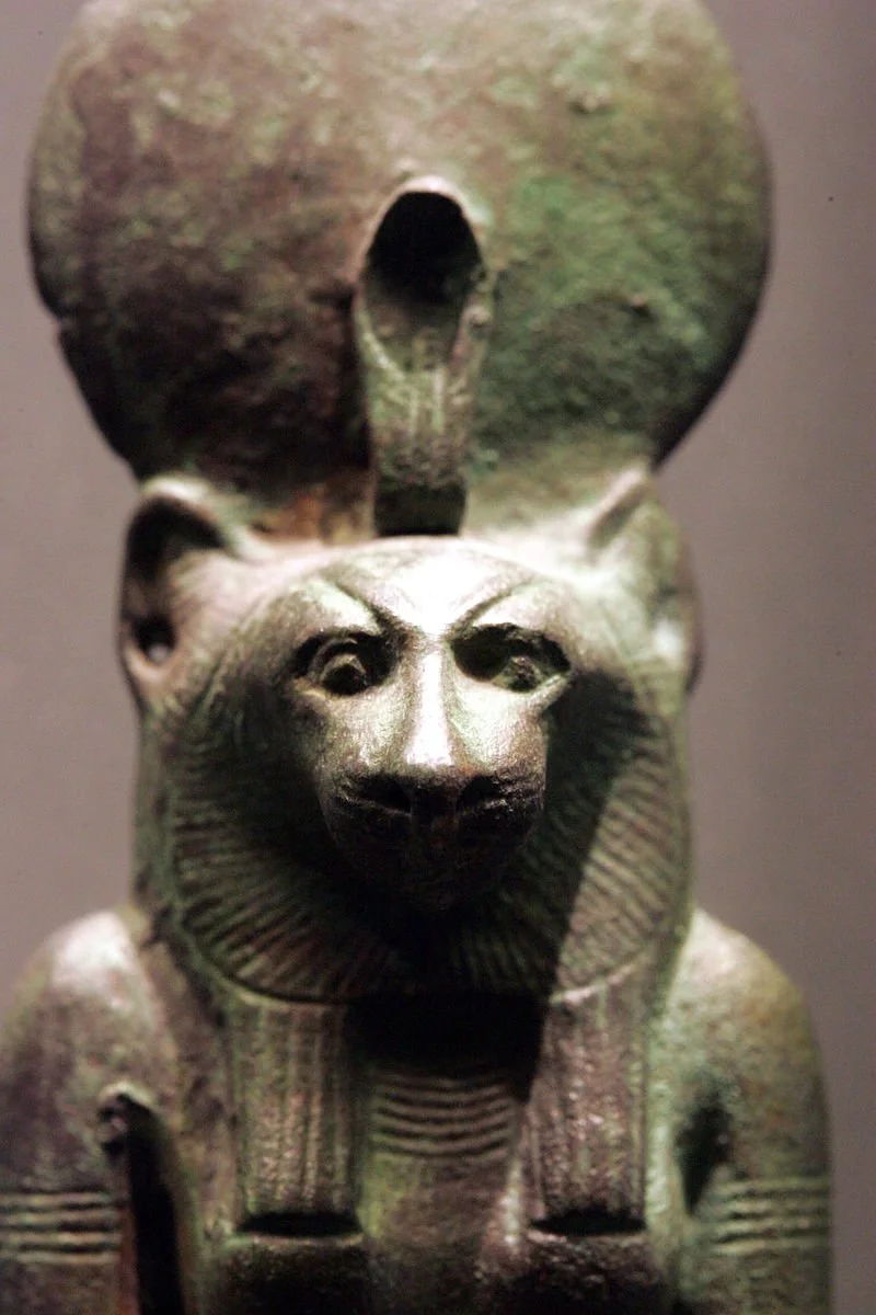 Ancient Egyptian sculpture of a lion with a human head, wearing a ceremonial collar, with a large, round object with a hole behind it.