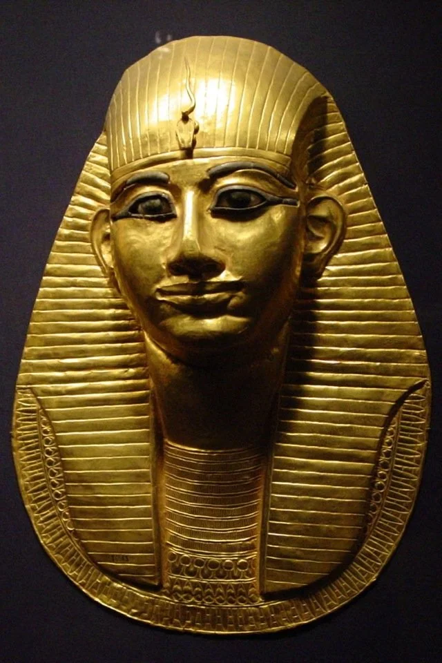 A golden funerary mask of ancient Egypt, depicting a smiling face with detailed headdress and necklace, set against a dark background.