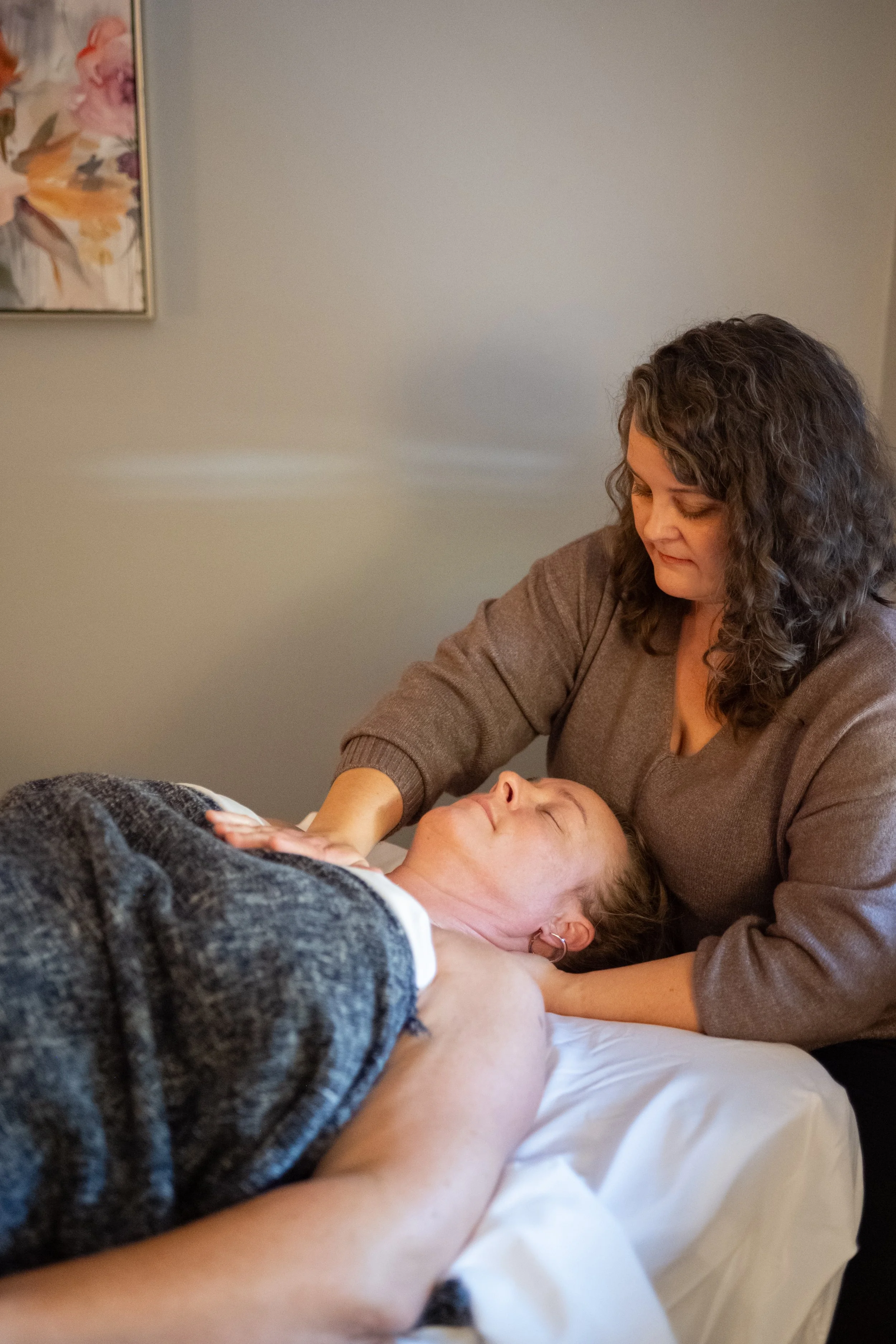 A woman receiving a massage, lying on her back with eyes closed, with another woman applying massage lotion or oil on her chest.