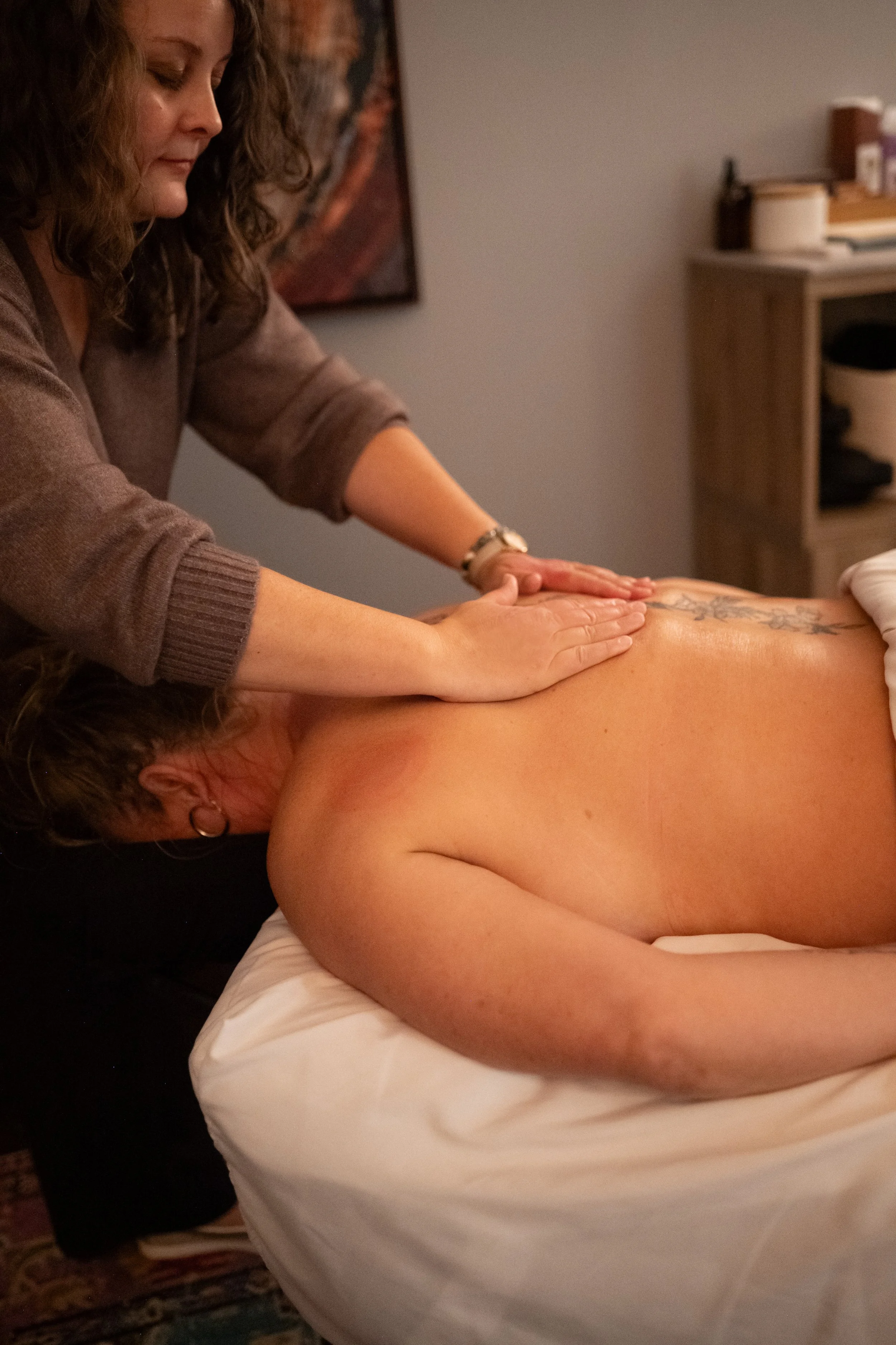 A woman giving a back massage to a person lying face down on a bed in a room with a pillow and shelf in the background.