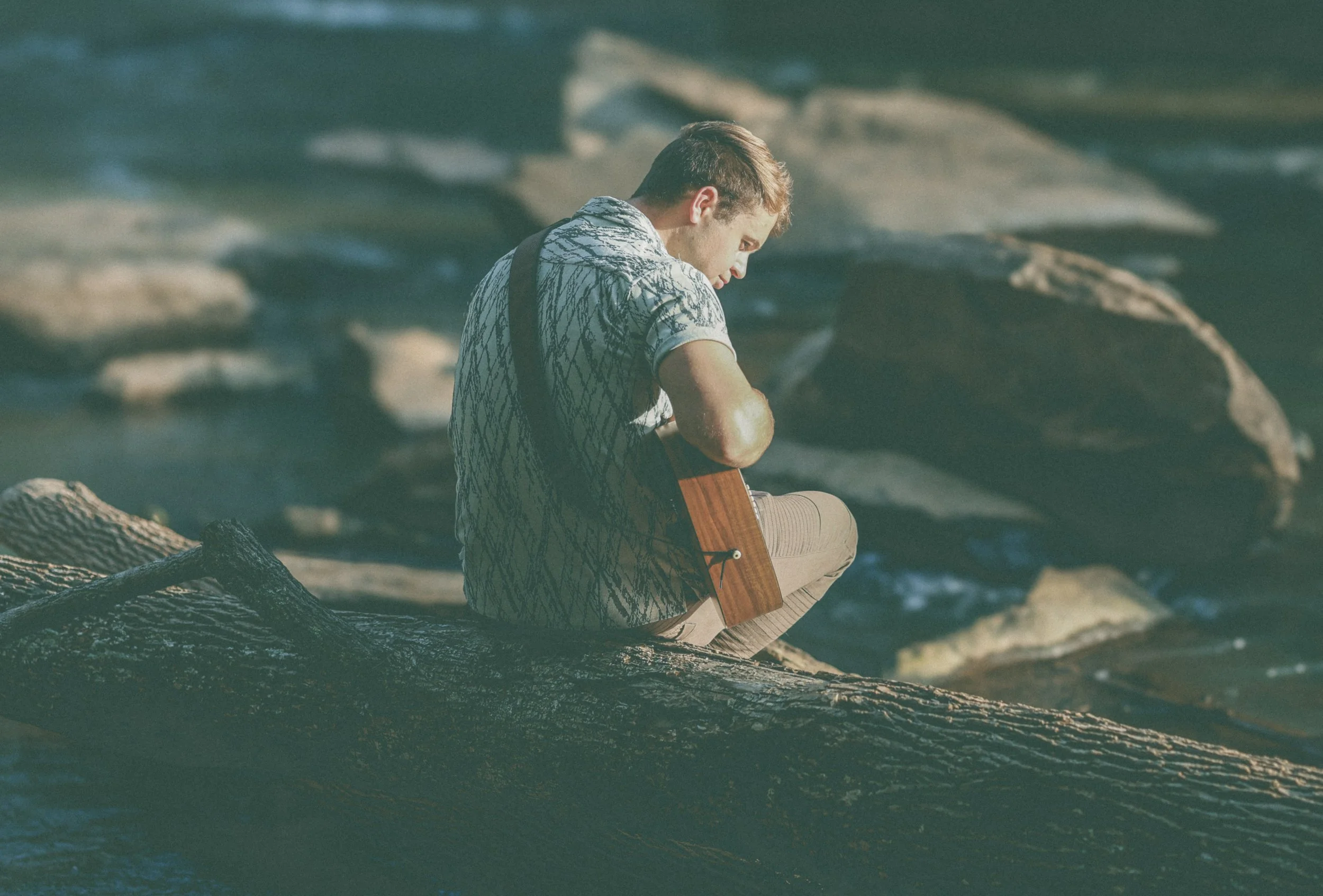 Jed Johnson sitting on rocks by a body of water, playing an acoustic guitar.