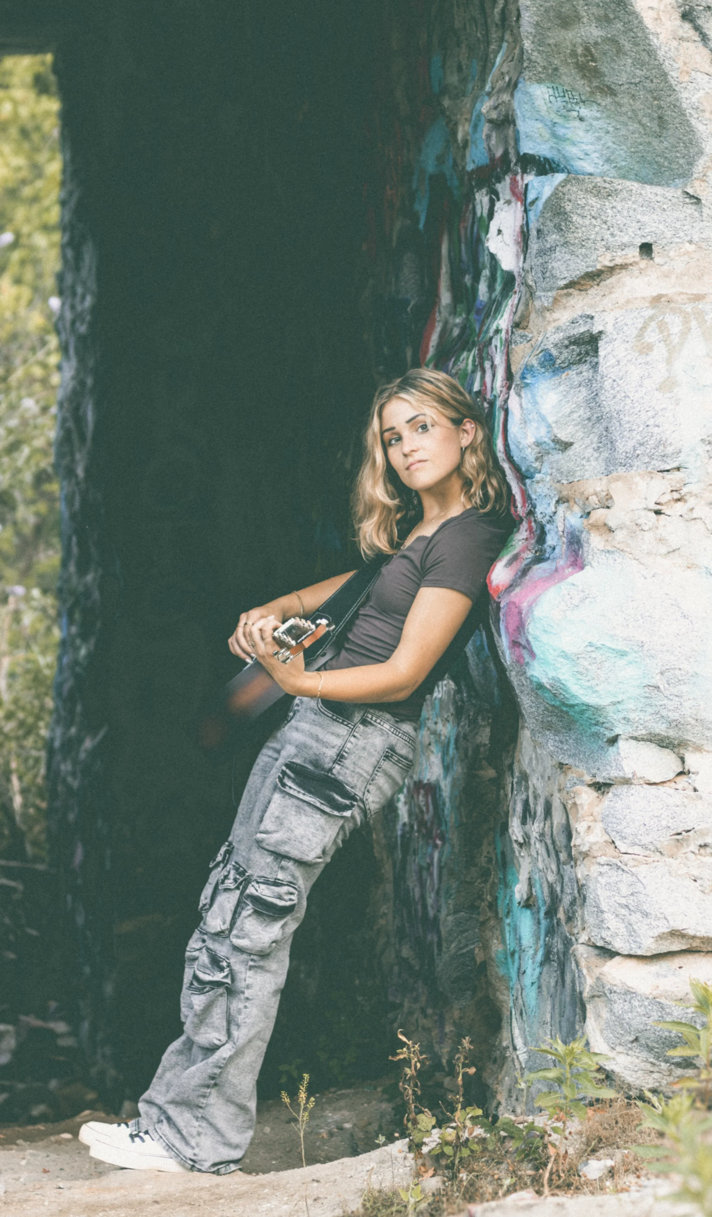 Erika McGonigal Johnson a young woman with blonde hair and a black t-shirt leaning against a colorful, graffiti-covered rock wall, holding a guitar. She is wearing baggy cargo pants and white sneakers, outdoors with greenery in the background.