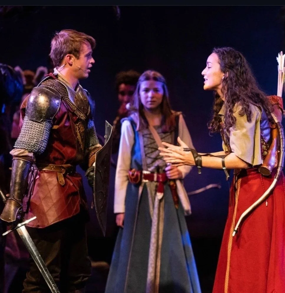 Three actors on stage in medieval costumes, two engaging in conversation, with a dark background and stage lighting. Jed Johnson plays Peter Pevensie in Prince Caspian. 