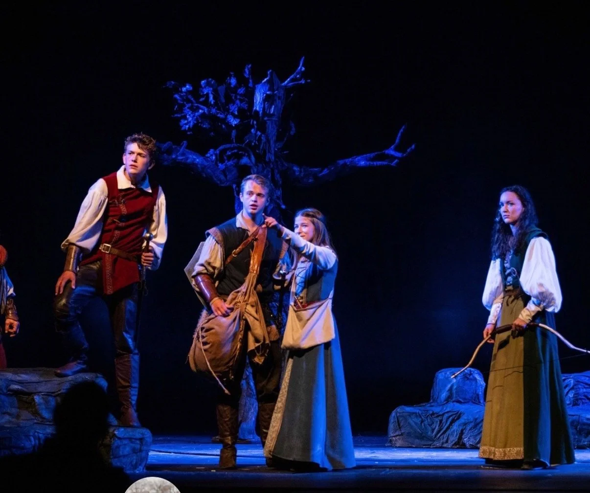 Theater scene with four young actors dressed in medieval or fantasy costumes, standing on a stage with a dark background and a large, twisted tree prop. Jed Johnson plays Peter Pevensie in Prince Caspian.