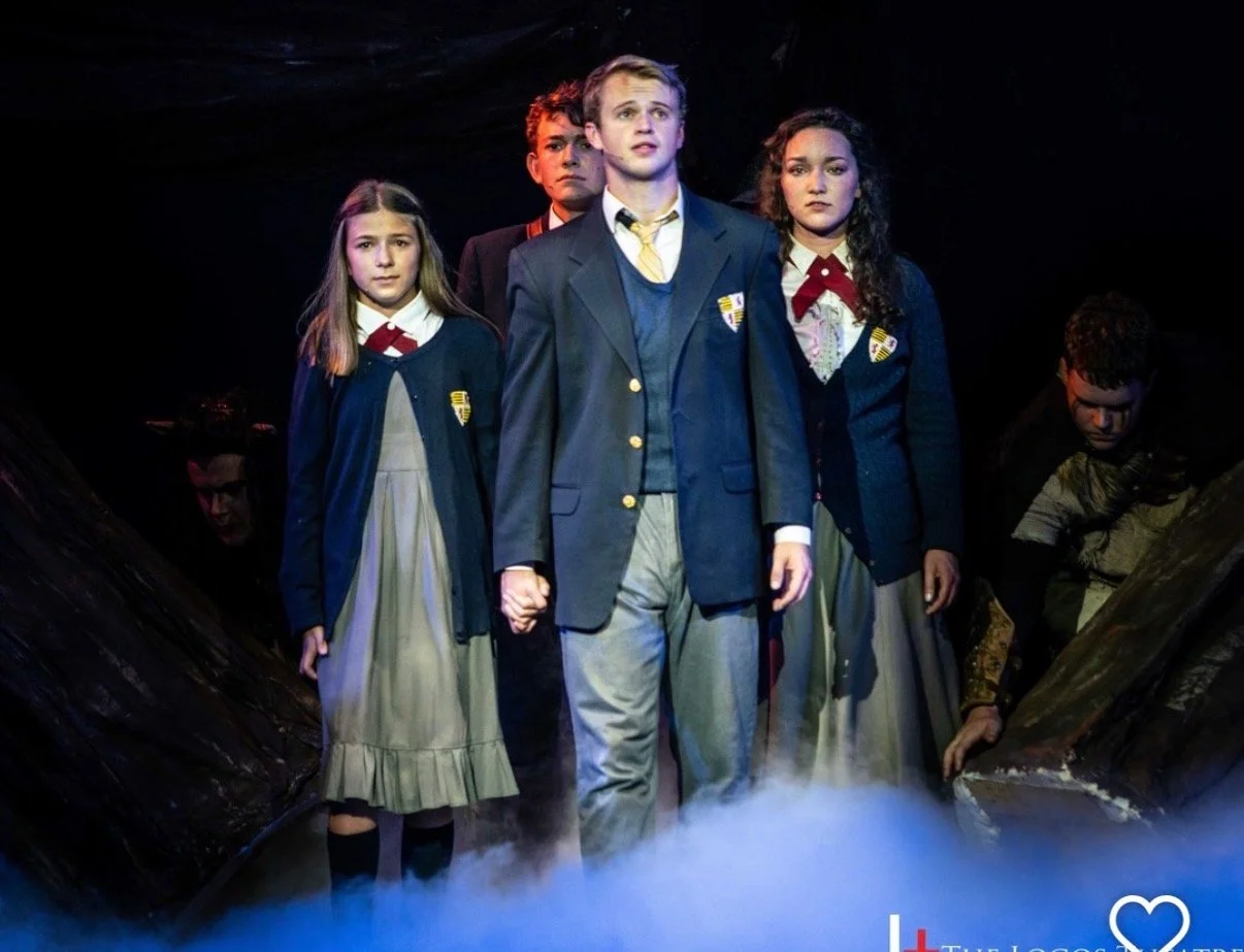 Four young actors wearing school uniforms standing on stage with serious expressions, surrounded by stage props and fog. Jed Johnson plays Peter Pevensie in Prince Caspian.