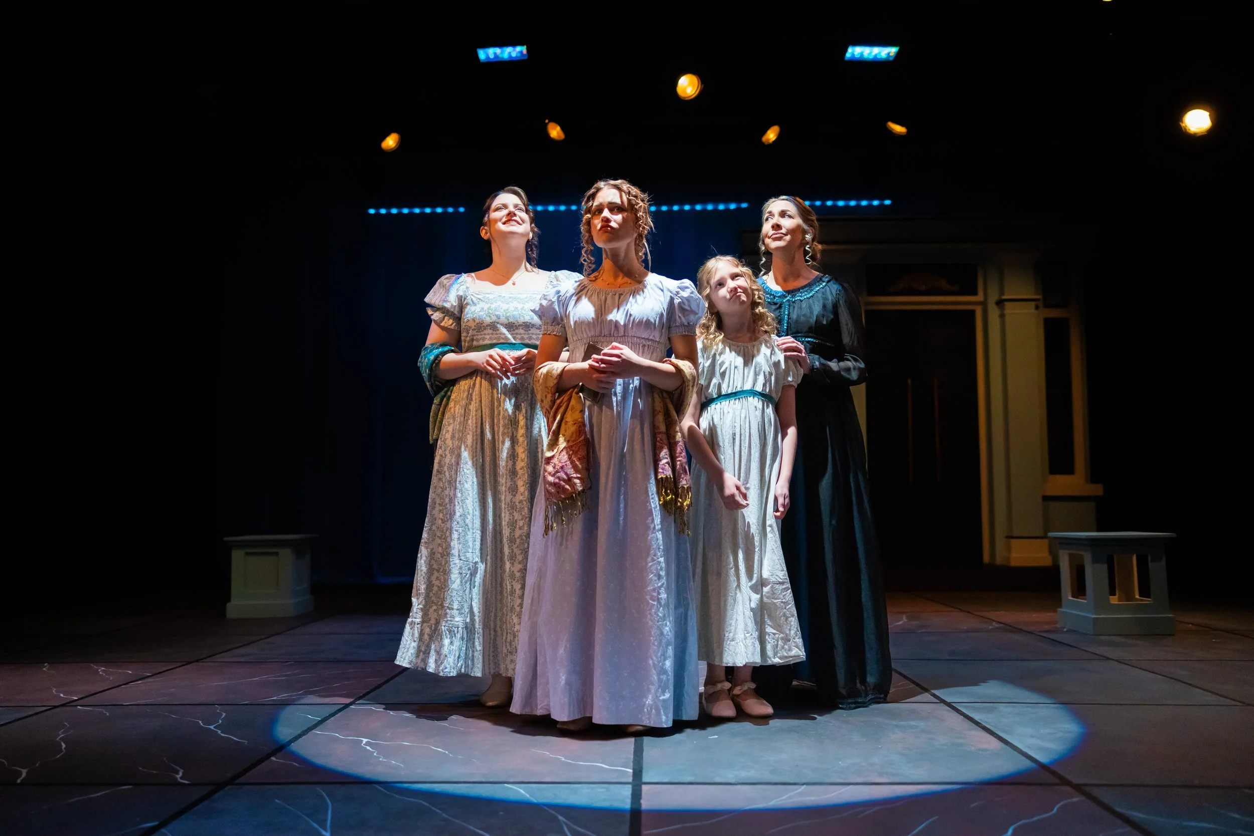 Four women in period costumes standing on a stage under yellow and blue stage lights.  Erika McGonigal Johnson plays Marianne Dashwood in Sense and Sensibility BJU 2025.
