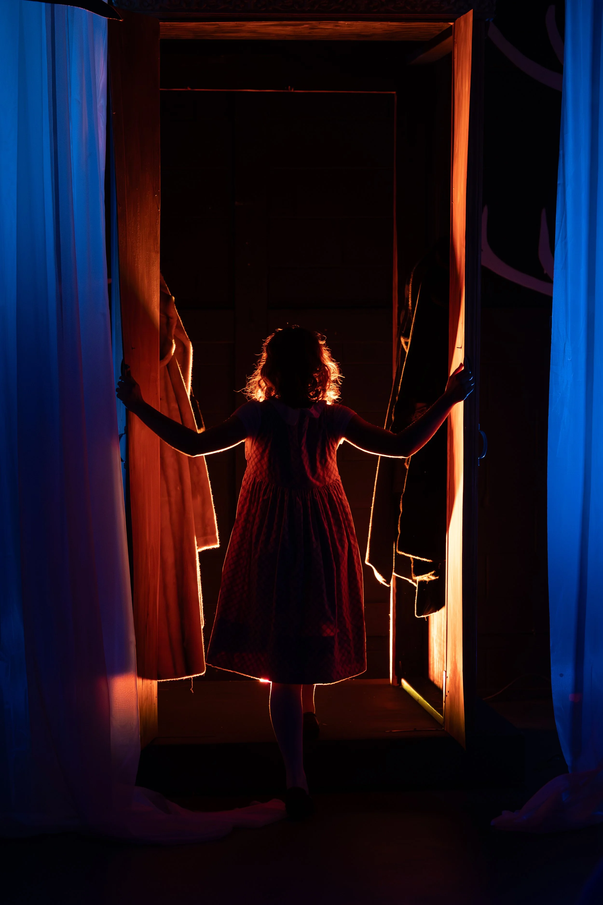 Silhouette of a girl in a dress standing in front of a dimly lit theater stage, holding open a curtain with bows on her hair. The stage is framed by curtains on either side and is glowing with warm, amber lighting. Erika McGonigal Johnson plays Lucy 