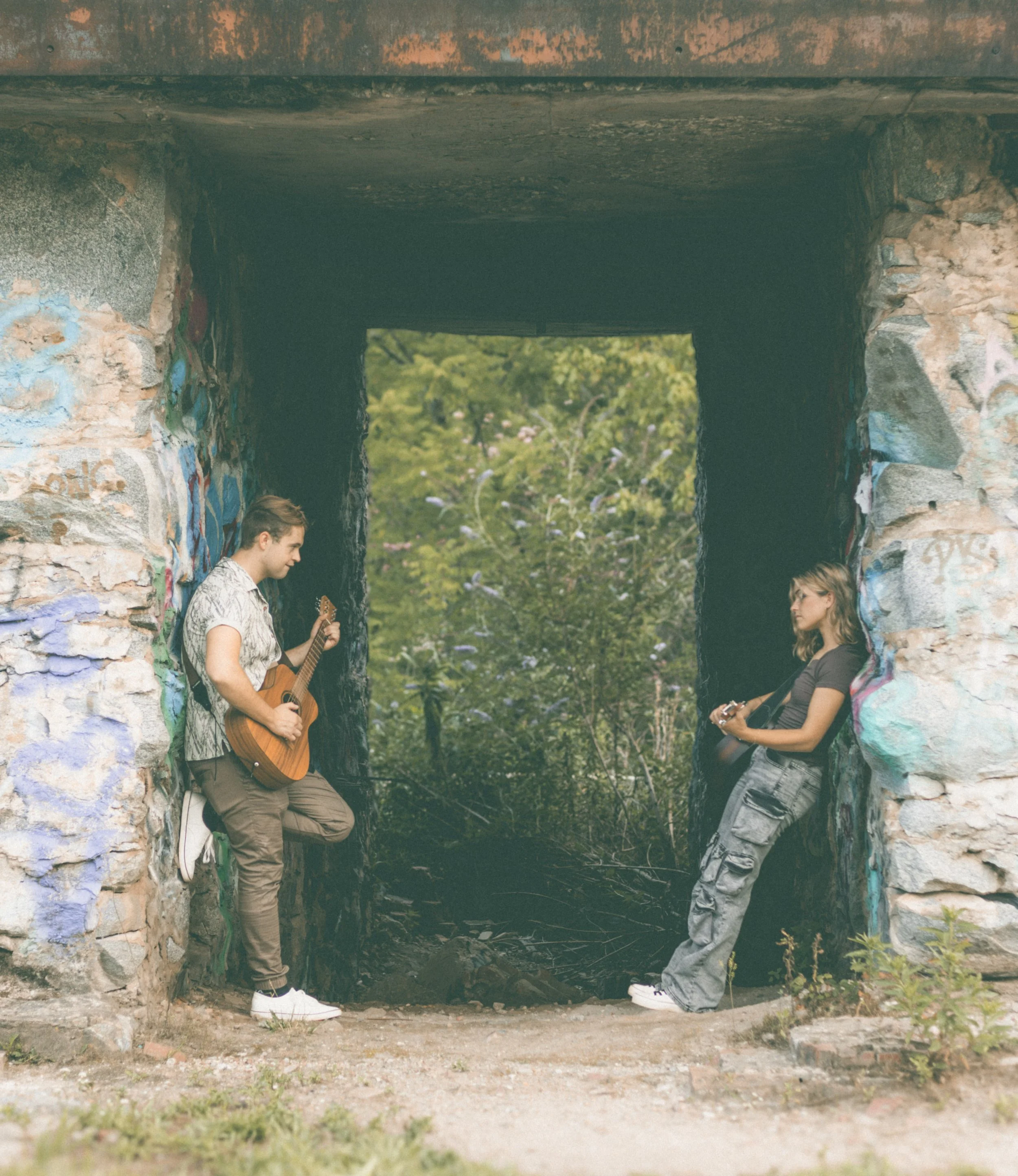 Erika McGonigal Johnson and Jed Johnson playing guitars in a graffiti-covered tunnel with trees visible through the opening